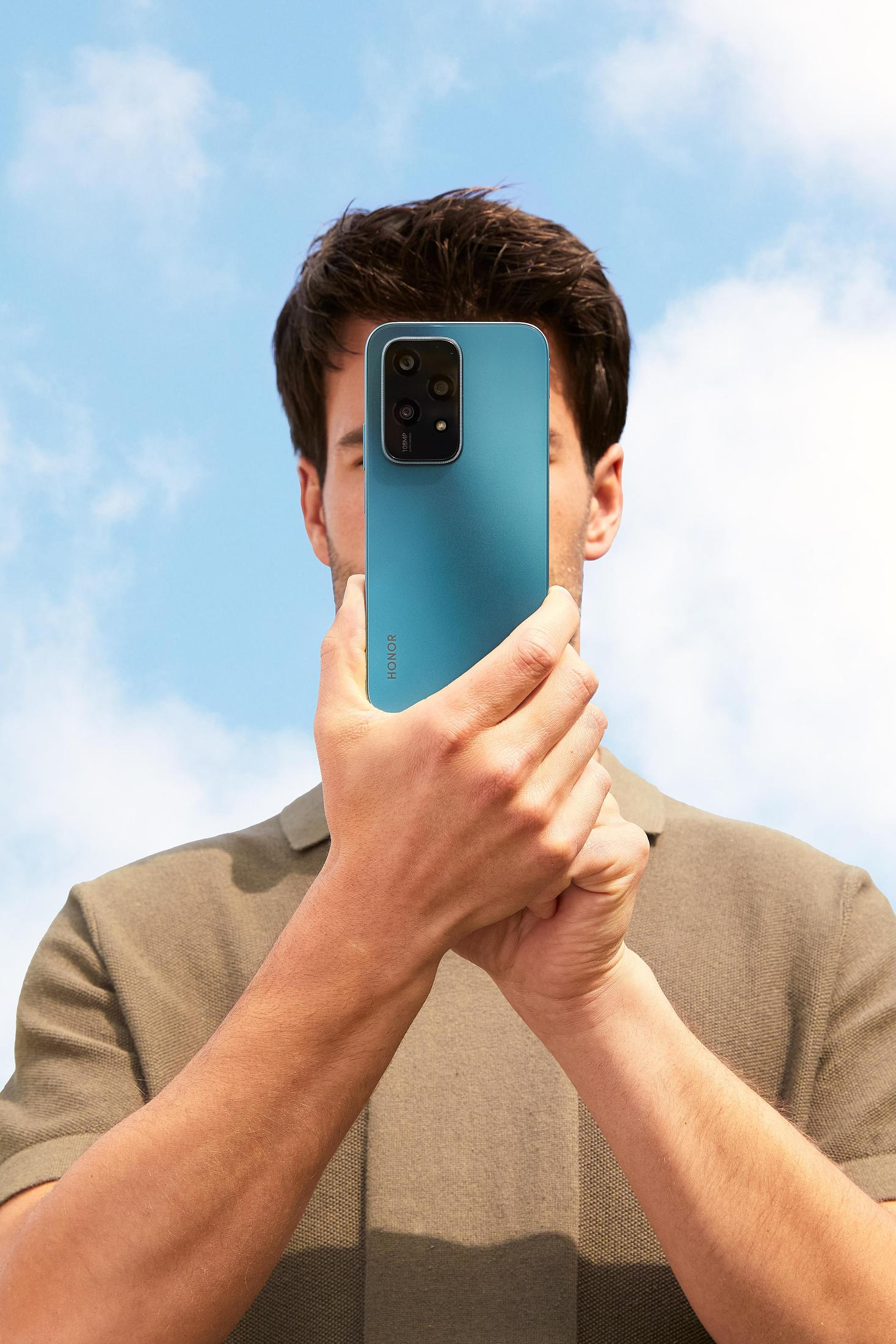 Eine Person hält ein tealfarbenes Telefon und verdeckt ihr Gesicht. Das Telefon hat Kameras und der Hintergrund ist ein blauer Himmel.