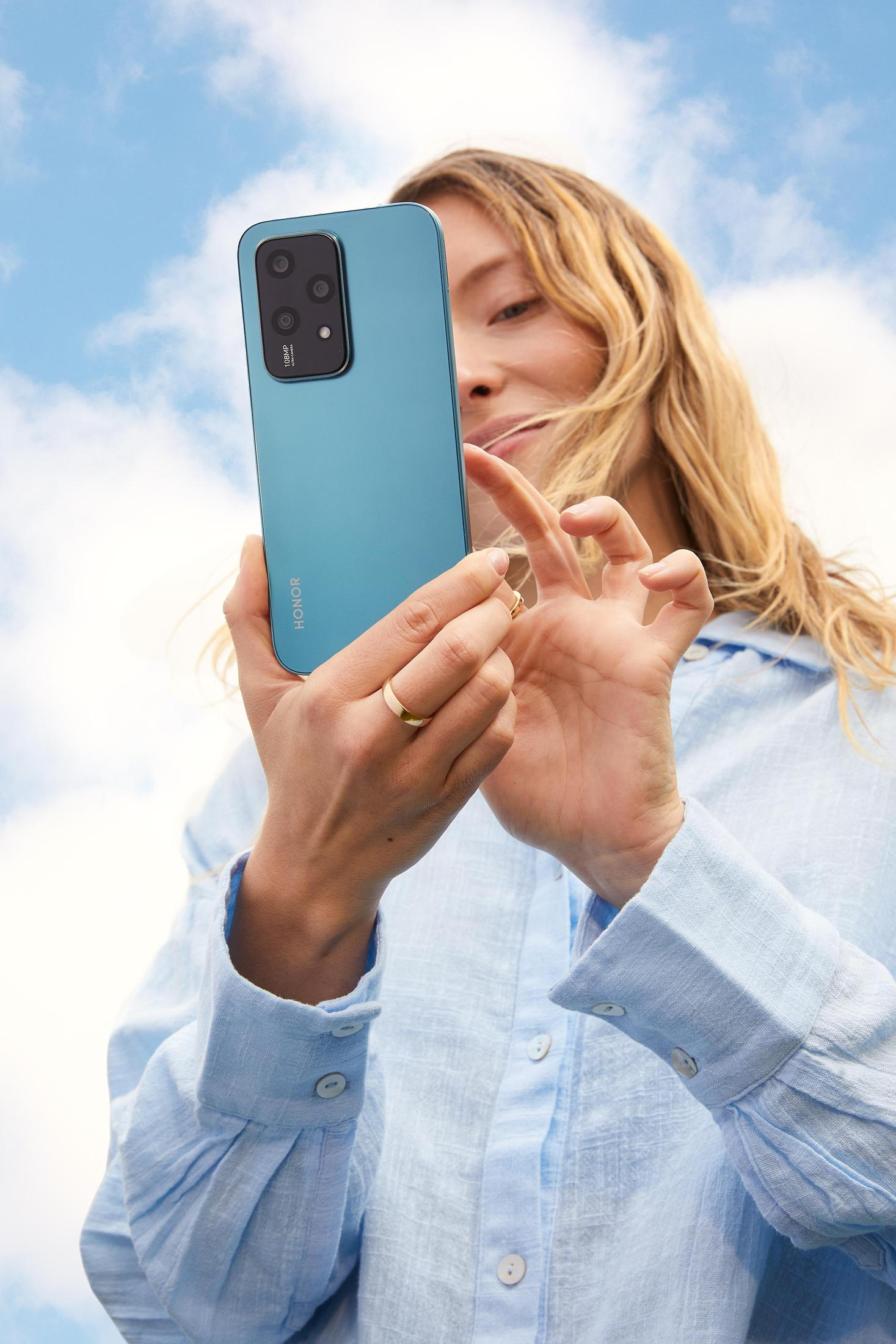 Frau hält tealfarbenes Telefon, blauer Himmel im Hintergrund. Sie trägt ein hellblaues Hemd und einen goldenen Ring.