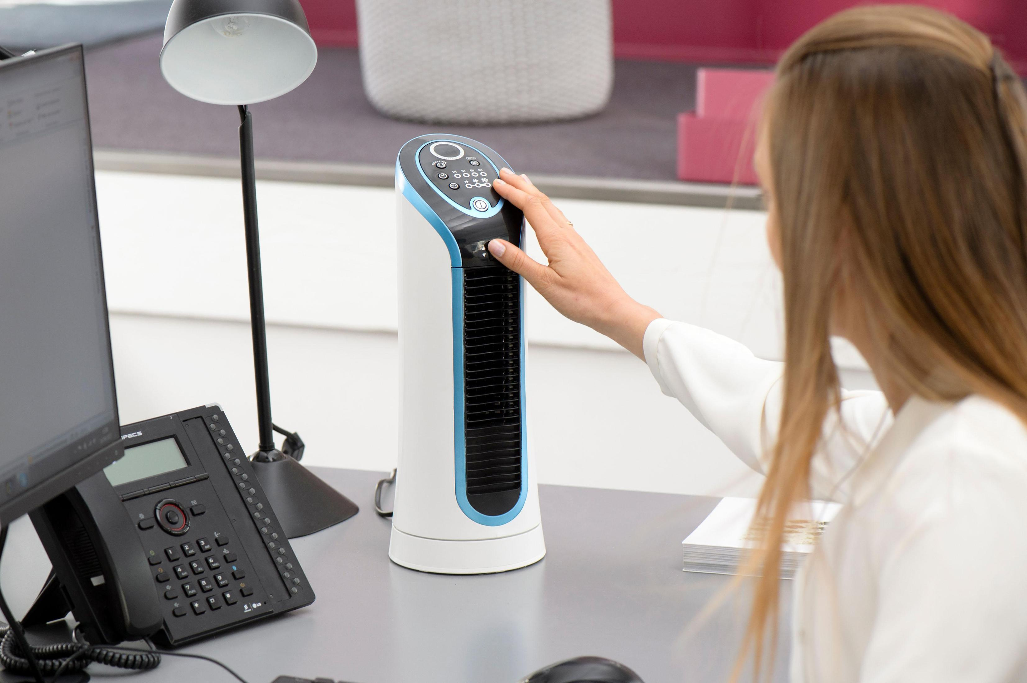 Dłoń kobiety na białym i niebieskim wentylatorze wieżowym na biurku. Widoczny telefon i lampa.