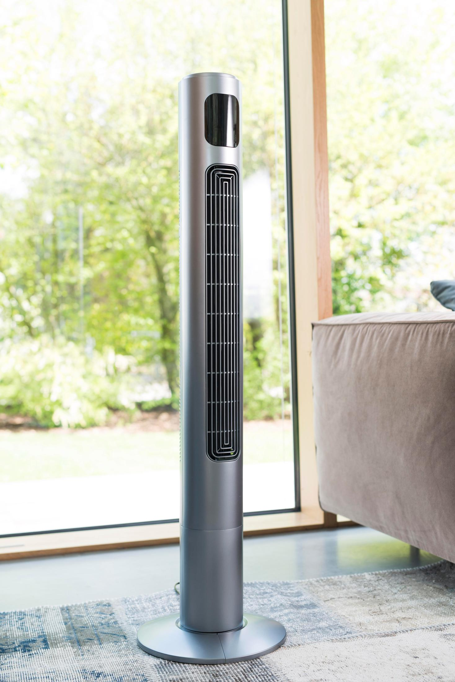 Silberner Turmventilator in einem Raum. Ein großes Fenster und eine Couch sind im Hintergrund.