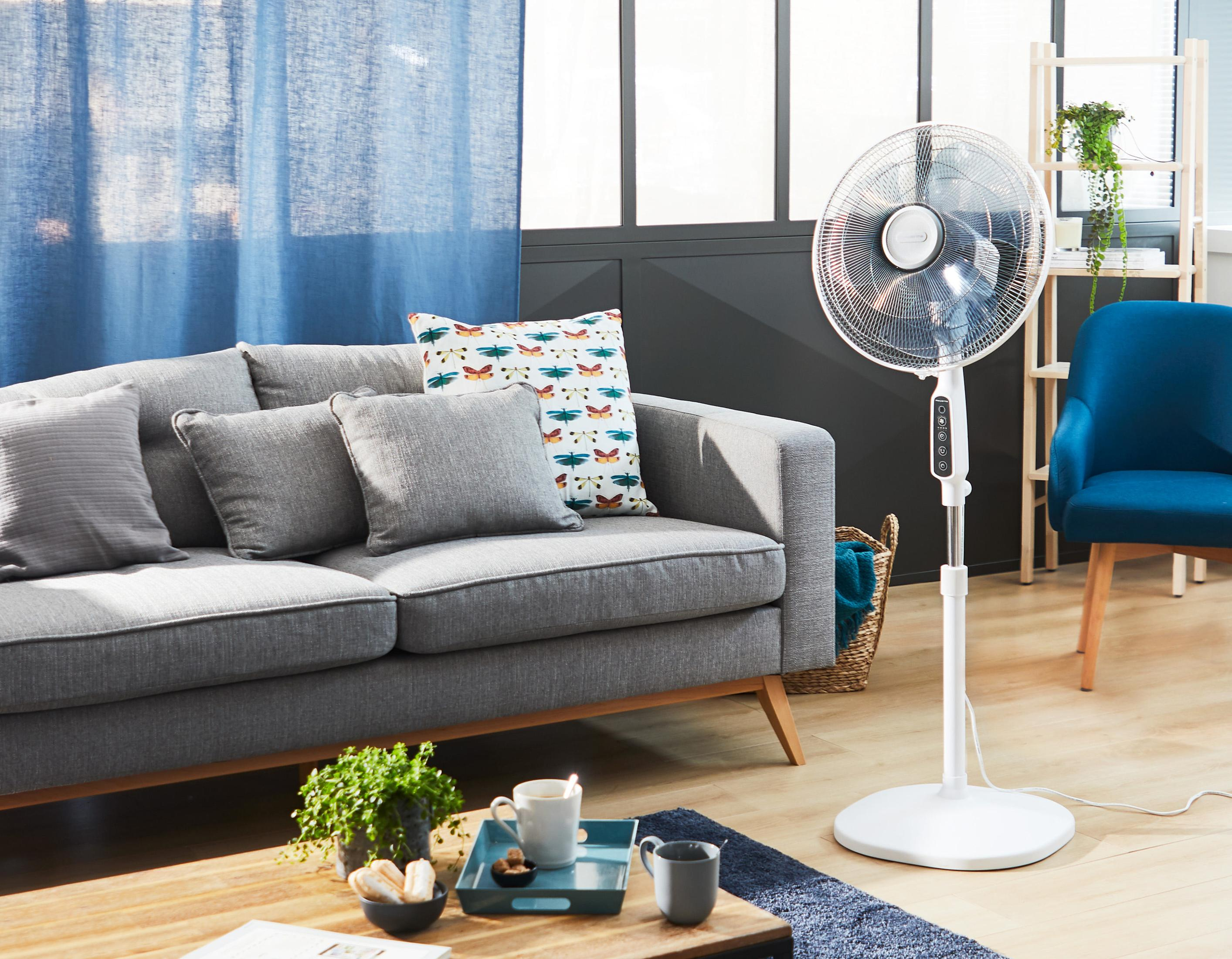 Scena w salonie z szarą sofą, wentylatorem i drewnianym stołem. Tło obejmuje okno.