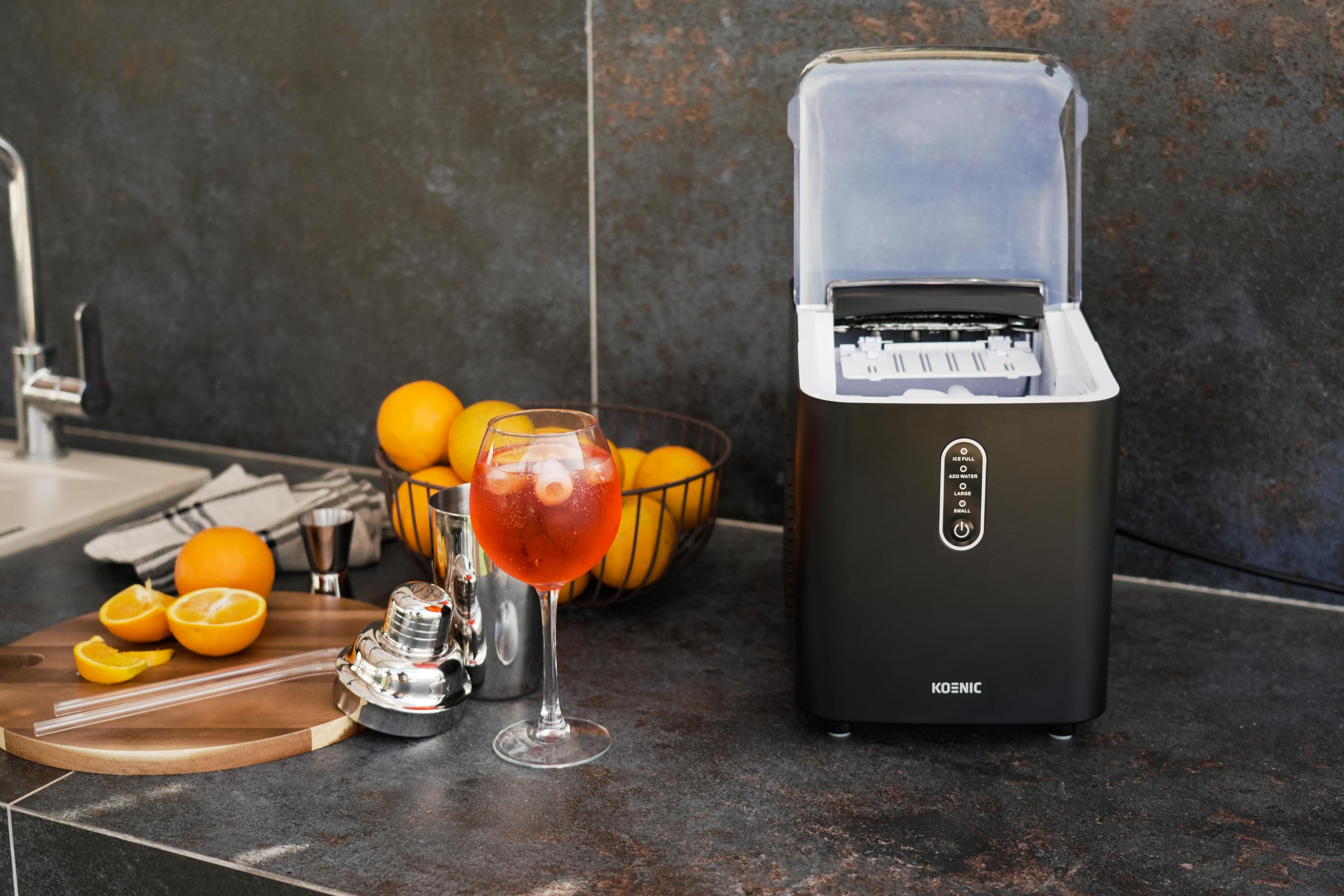 Eismaschine mit offenem Deckel, Glas mit Getränk, Orangen und Cocktailshaker auf dunkler Oberfläche.