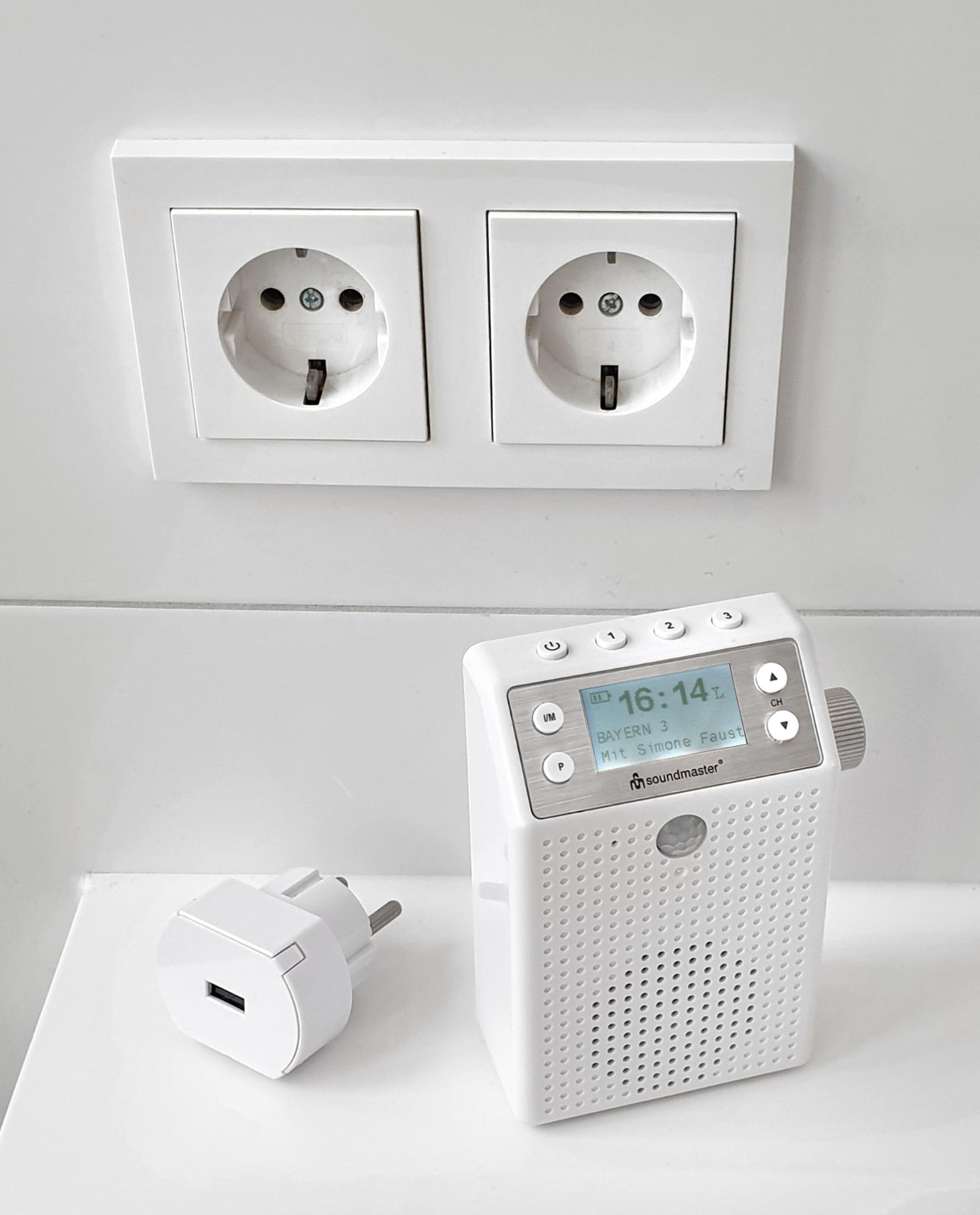 Weißes Radio und Stecker neben einer Doppelsteckdose. Radio zeigt 16:14 und Sender BAYERN 3.
