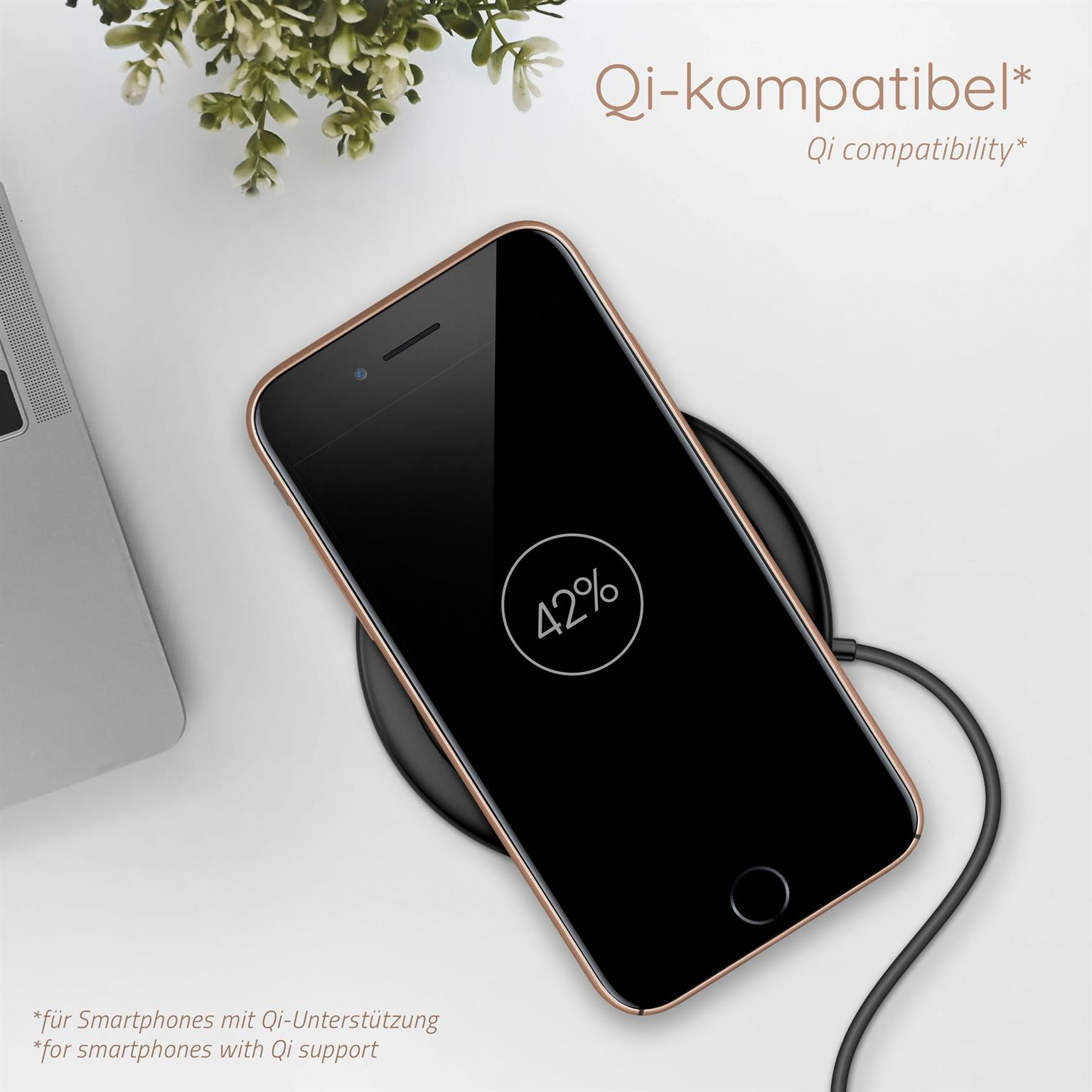Ein Telefon auf einem kabellosen Ladegerät zeigt 42 % Ladung an. Ein Laptop und eine Pflanze sind im Hintergrund.