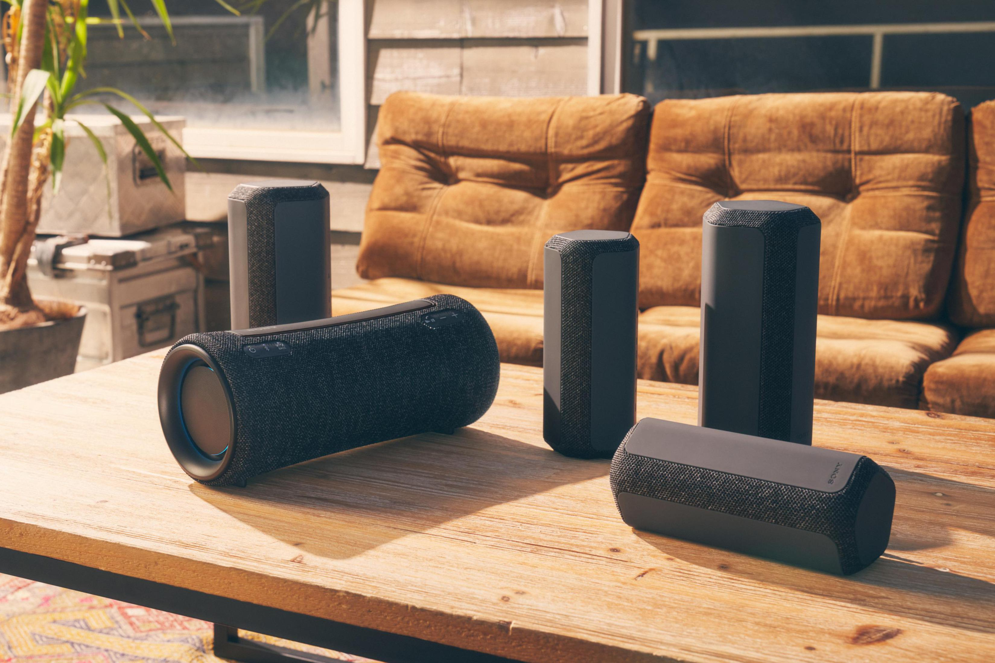 Múltiples altavoces negros en una mesa de madera. Cojines y plantas en el fondo.