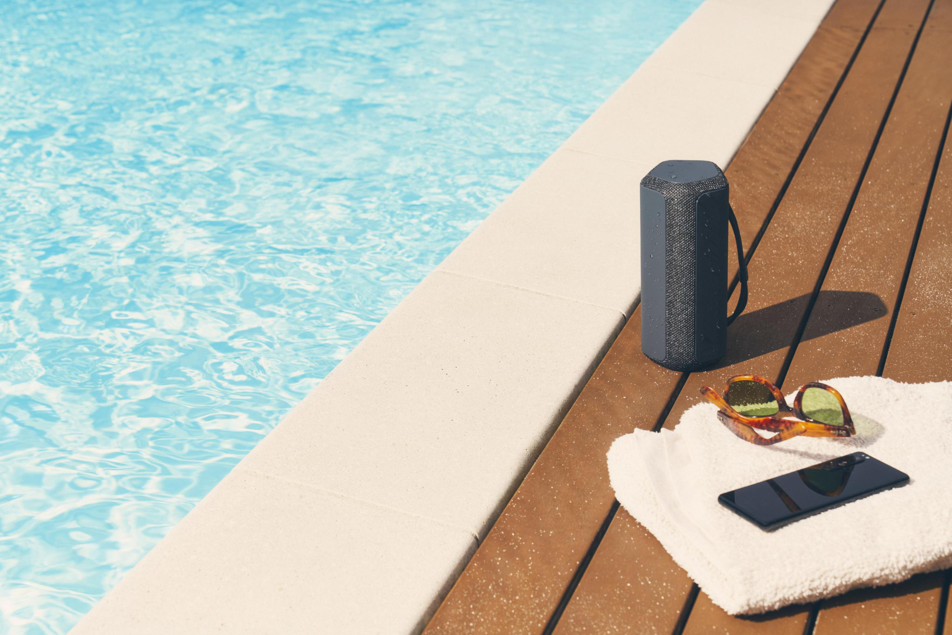 Bluetooth-Lautsprecher und Sonnenbrille auf einem Handtuch am Pool. Wasser und Holzdeck im Hintergrund.