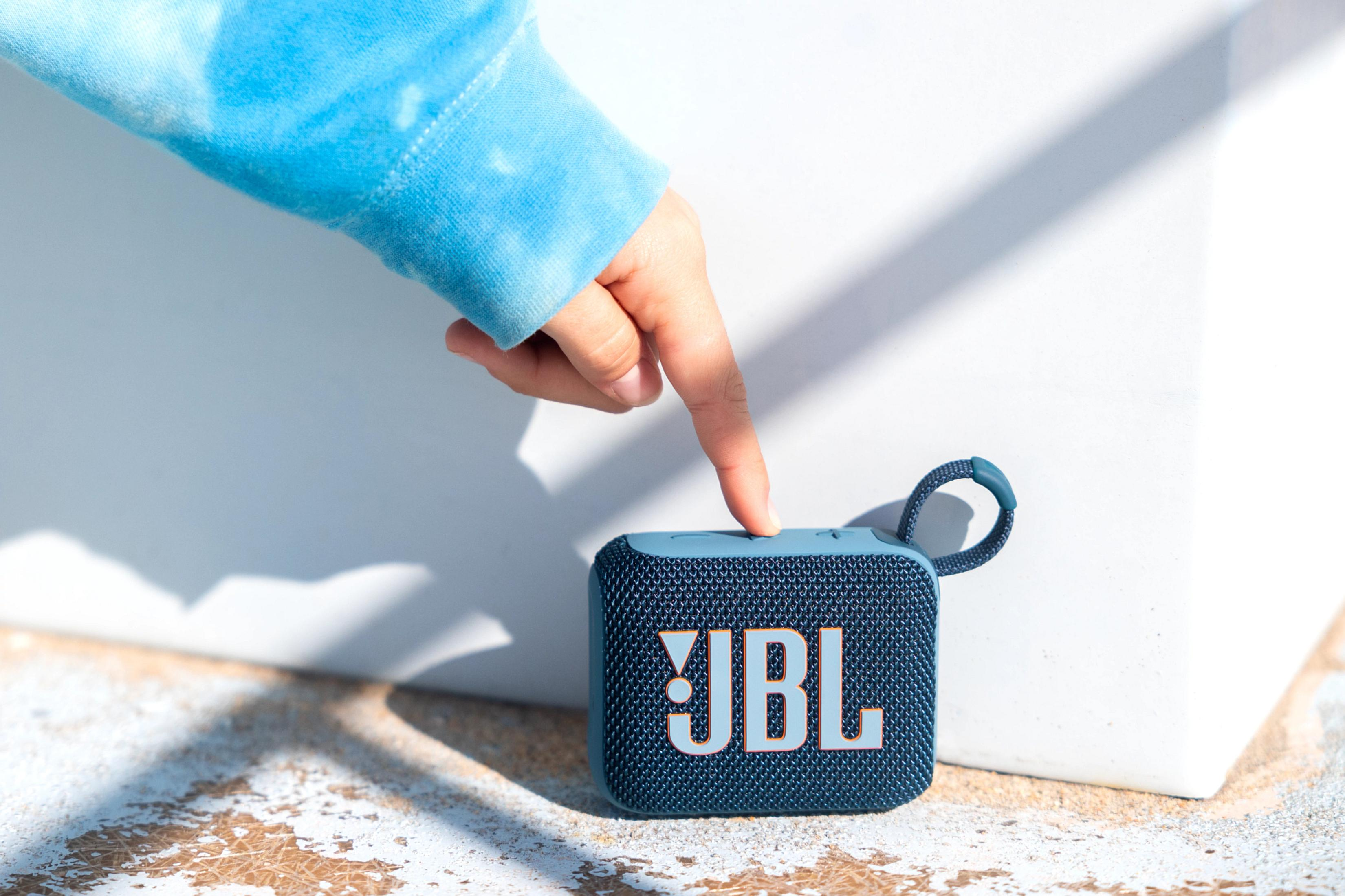 Bir el, JBL logosunun net bir şekilde görülebildiği, küçük mavi bir JBL hoparlör üzerindeki bir düğmeye basıyor.