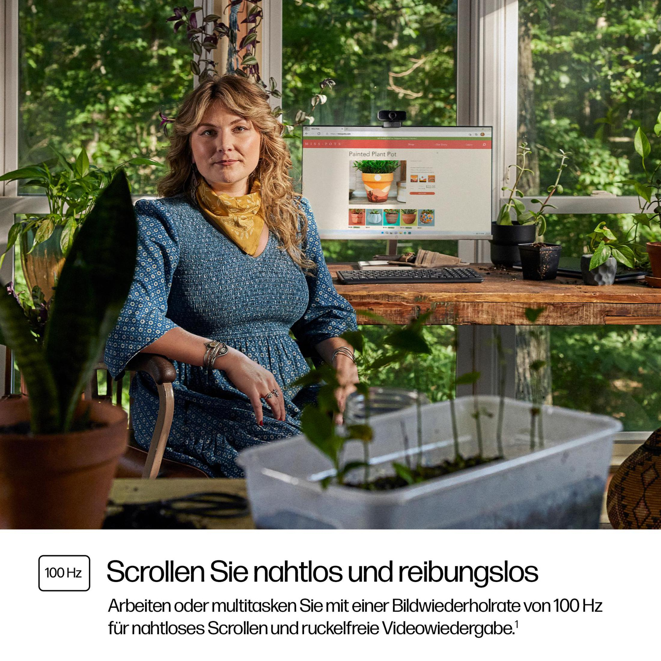 Kobieta w niebieskiej sukience przy biurku z roślinami, używająca komputera z kamerką.