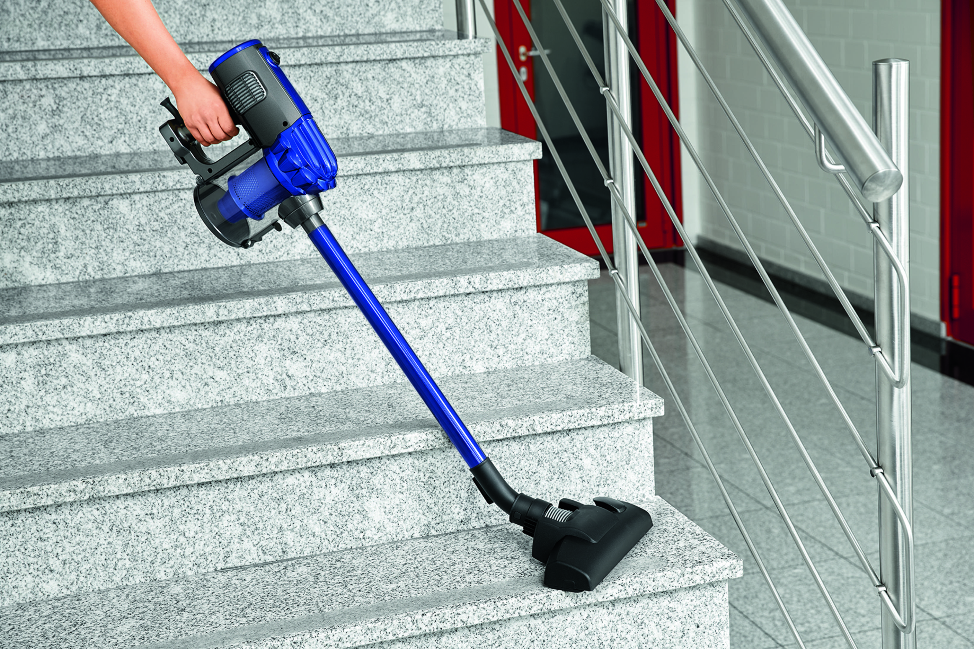 Osoba odkurza schody niebiesko-czarnym odkurzaczem bezprzewodowym.