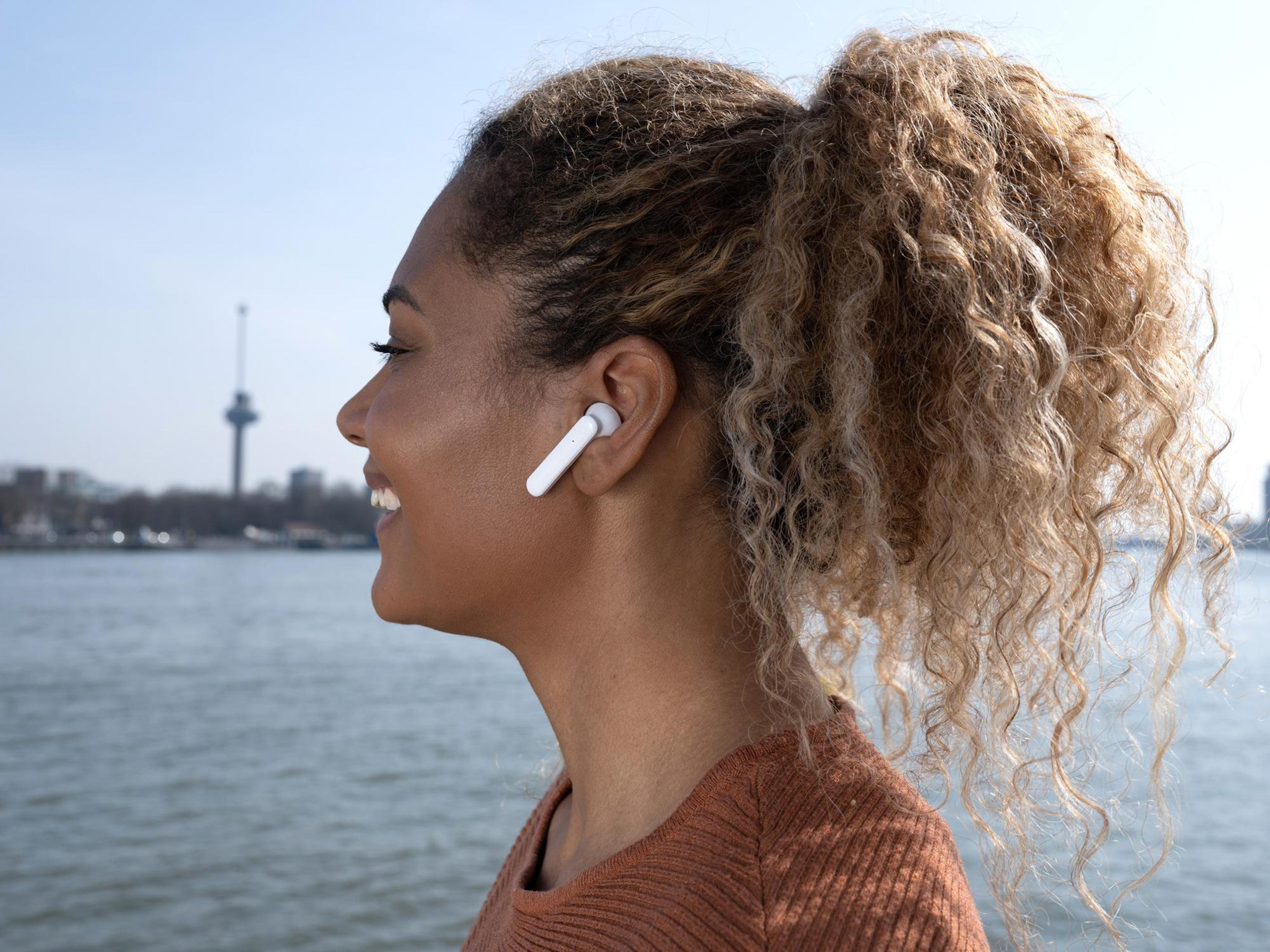 Kobieta z kręconymi włosami nosi białą słuchawkę, uśmiechając się w pobliżu wody z budynkami.