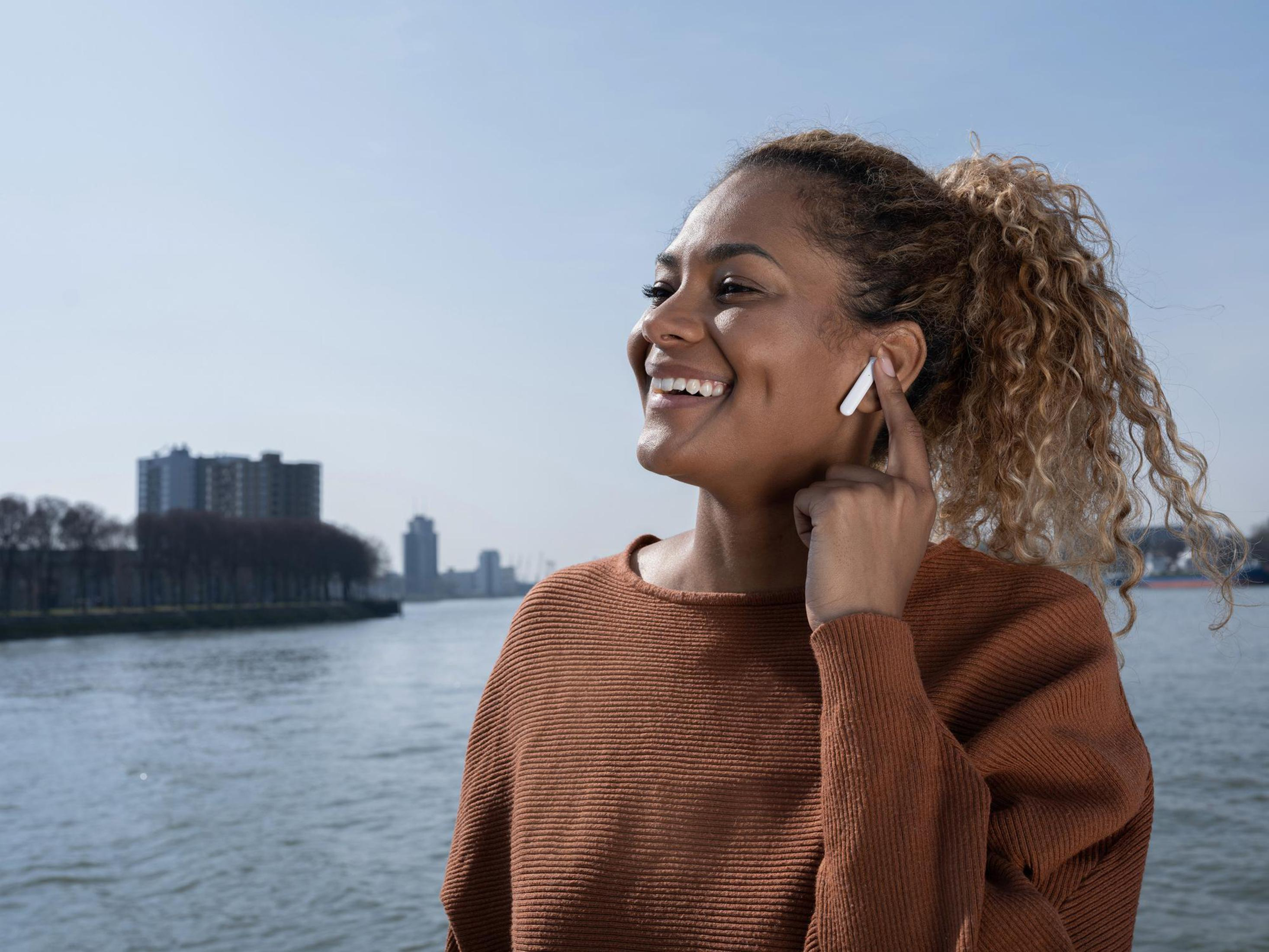 Kobieta ze słuchawkami uśmiecha się nad wodą. Panorama miasta w tle. Słoneczny dzień.