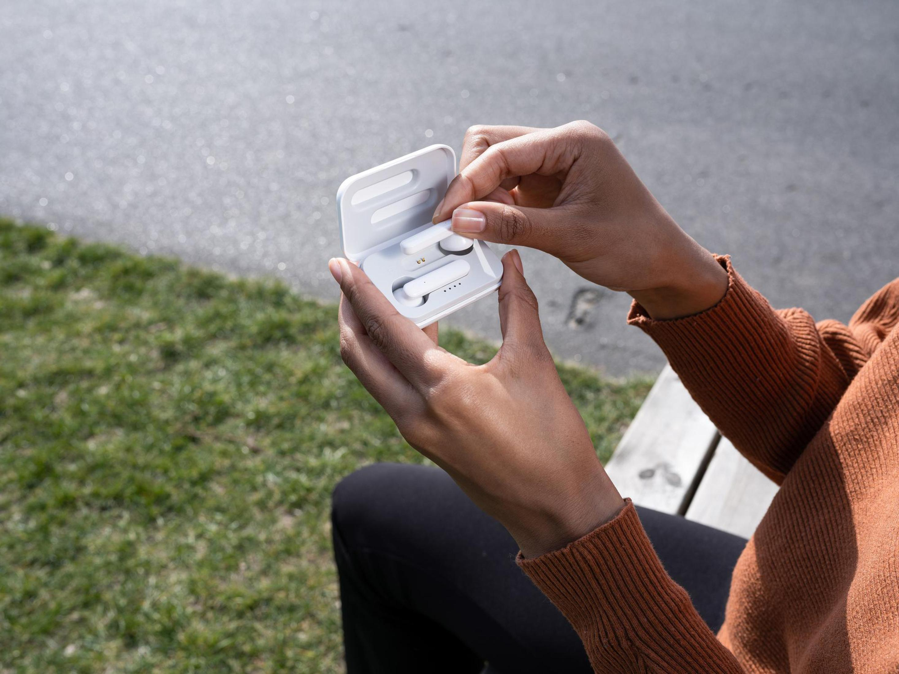 Osoba trzymająca białe etui, otwierająca je, aby odsłonić dwie białe słuchawki douszne w środku. Zielone tło trawy.