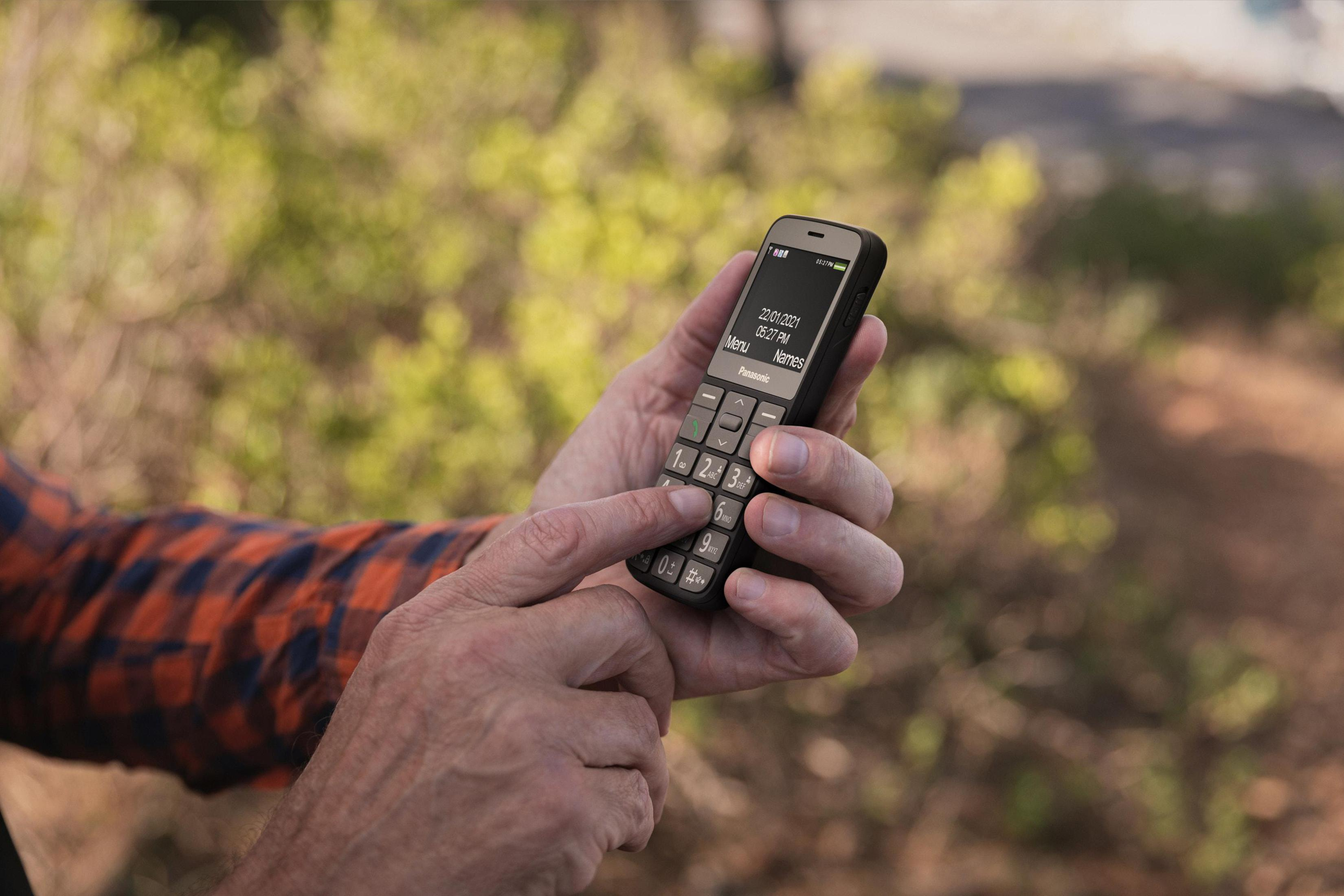 Hände einer Person, die ein kleines schwarzes Panasonic-Telefon halten, wobei der Daumen eine Taste drückt.