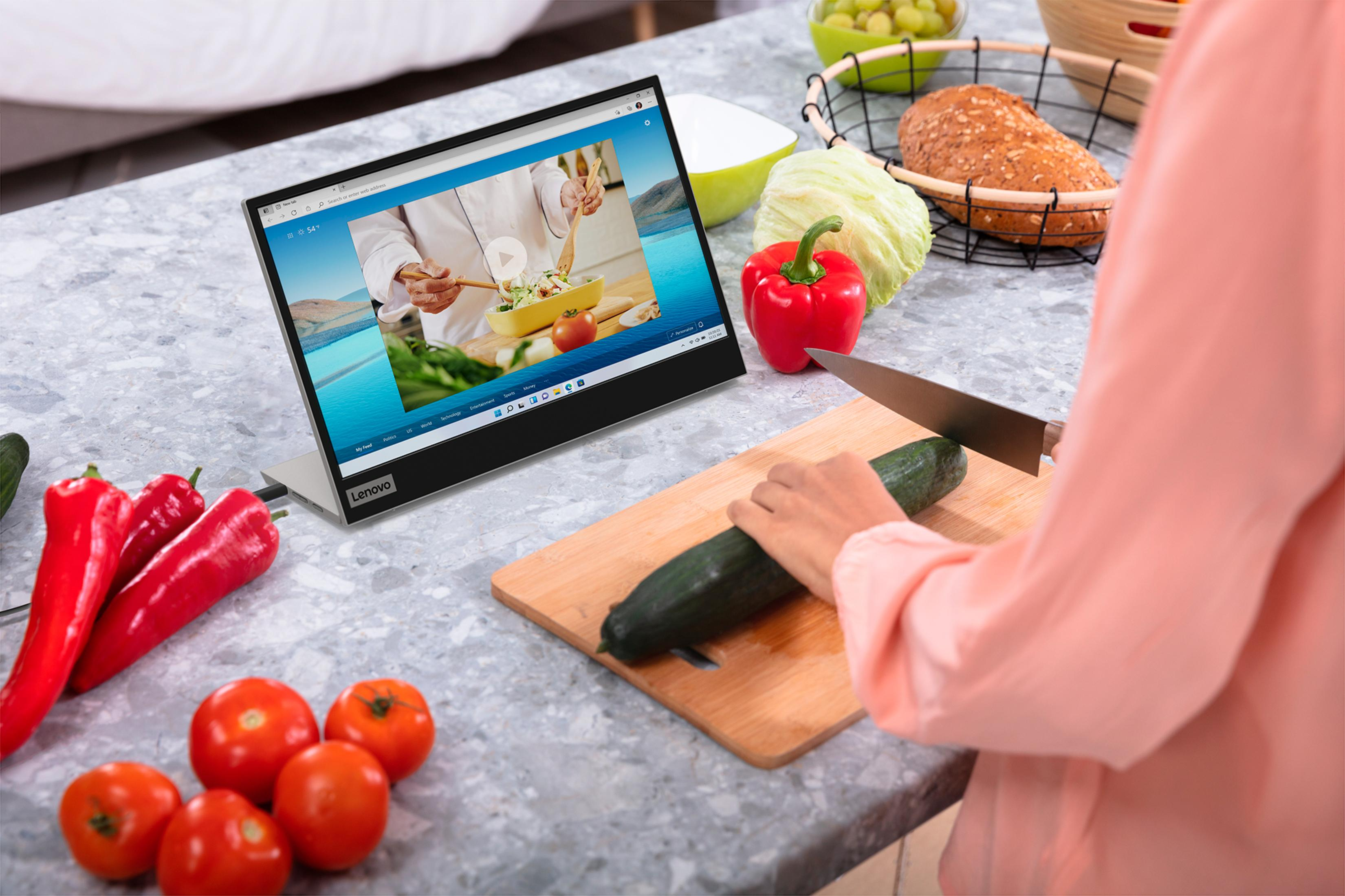 Osoba krojąca ogórek obok laptopa odtwarzającego film z gotowania, z pomidorami i papryką.