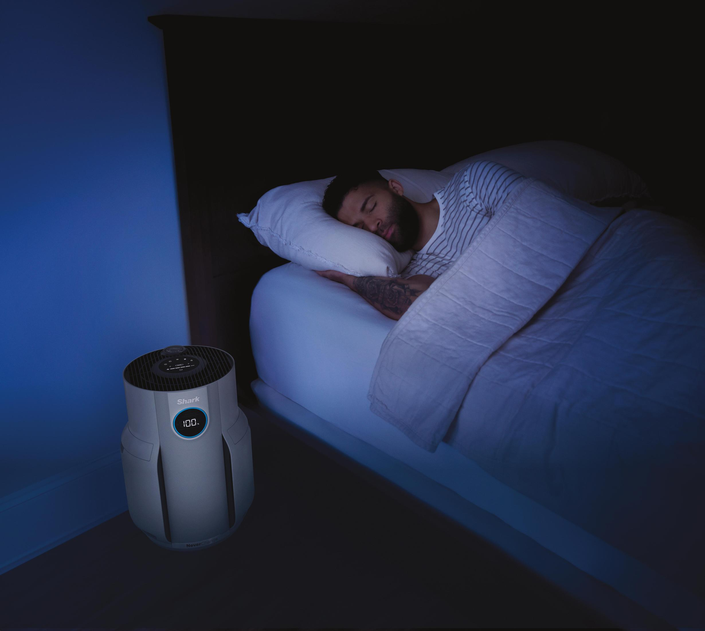 Ein Mann schläft in einem Bett mit einem Luftreiniger daneben. Der Luftreiniger zeigt 100% auf seinem Display.