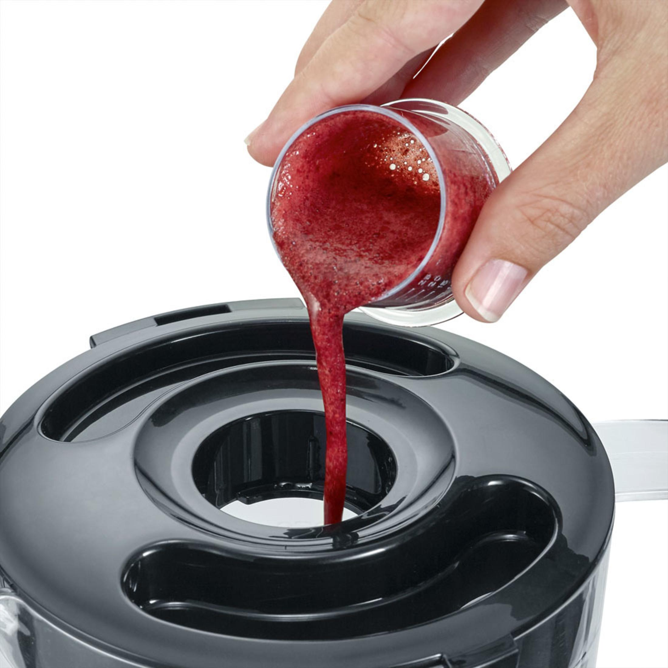 Dłoń wlewa czerwone smoothie do blendera. Szklany pojemnik trzymany jest nad blenderem.