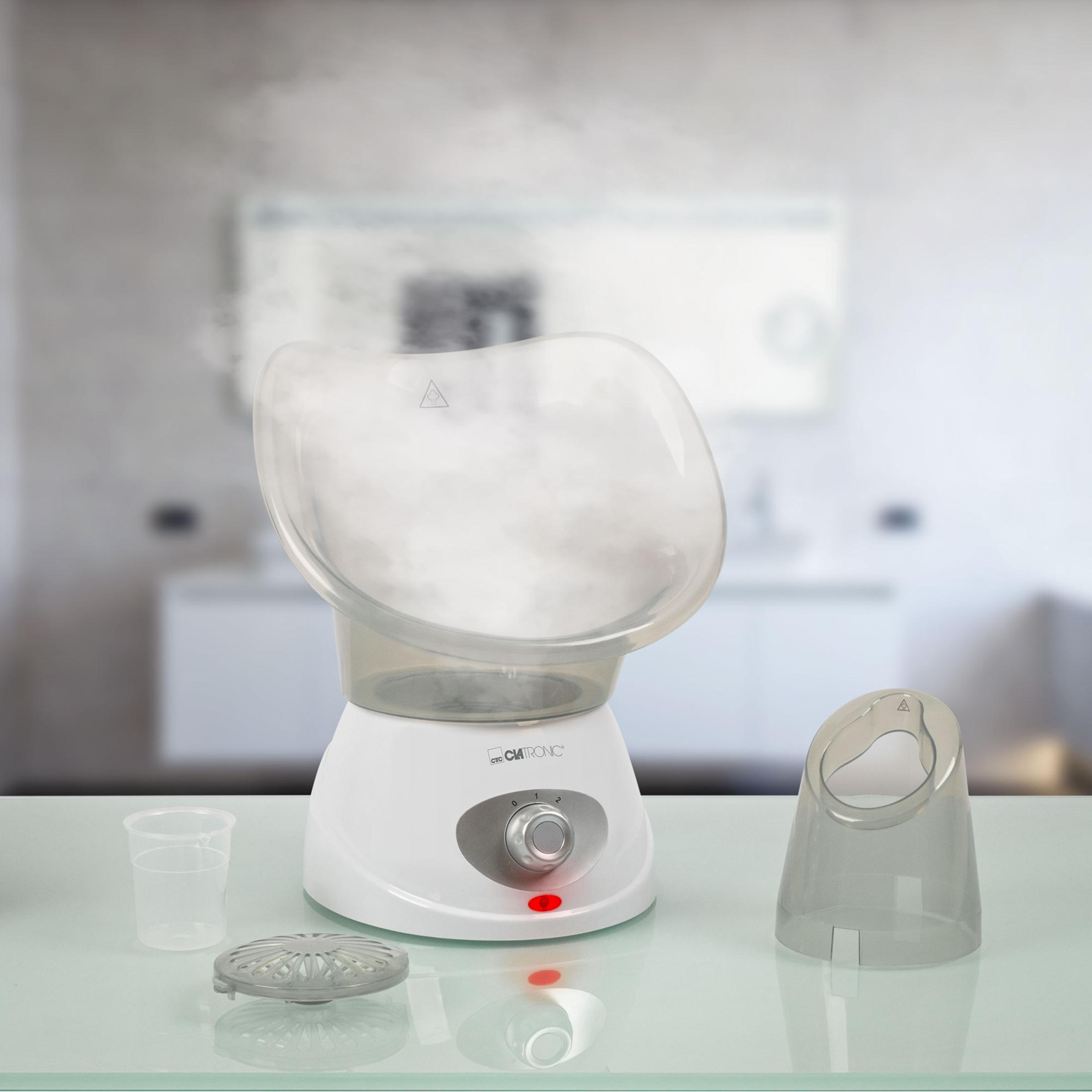 Parownica do twarzy z parą, kubkiem na wodę i dodatkowym elementem na białym stole.