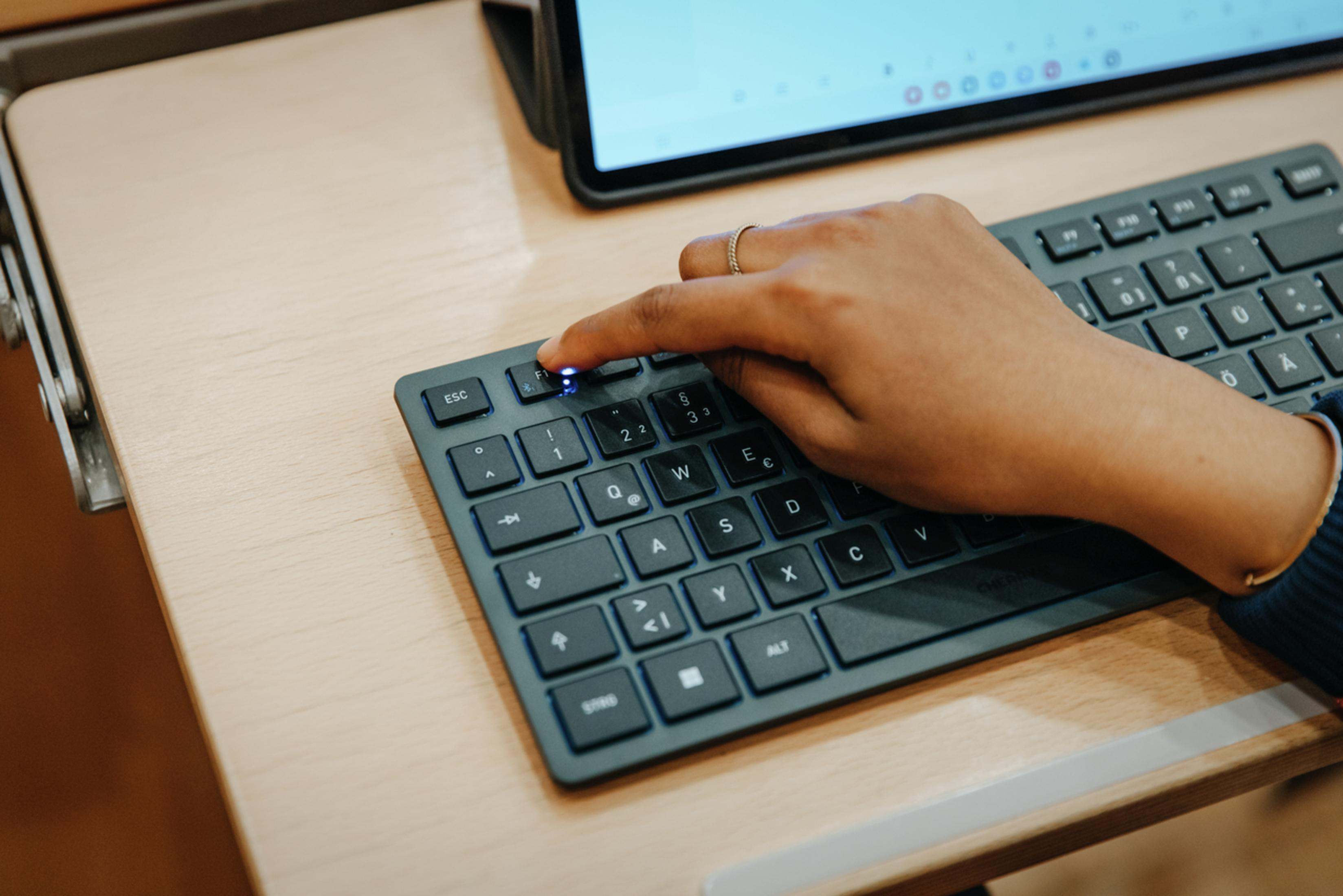 Main appuyant sur le bouton d'alimentation d'un clavier noir, tablette au-dessus. Bureau en bois beige.