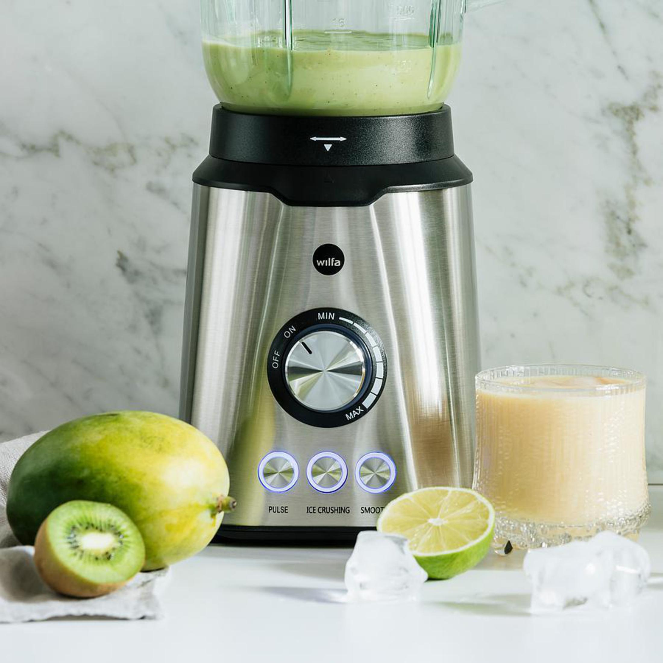 Mixer mit grünem Smoothie. Der Mixer hat einen silbernen Sockel und ein Glas. Zutaten drumherum.