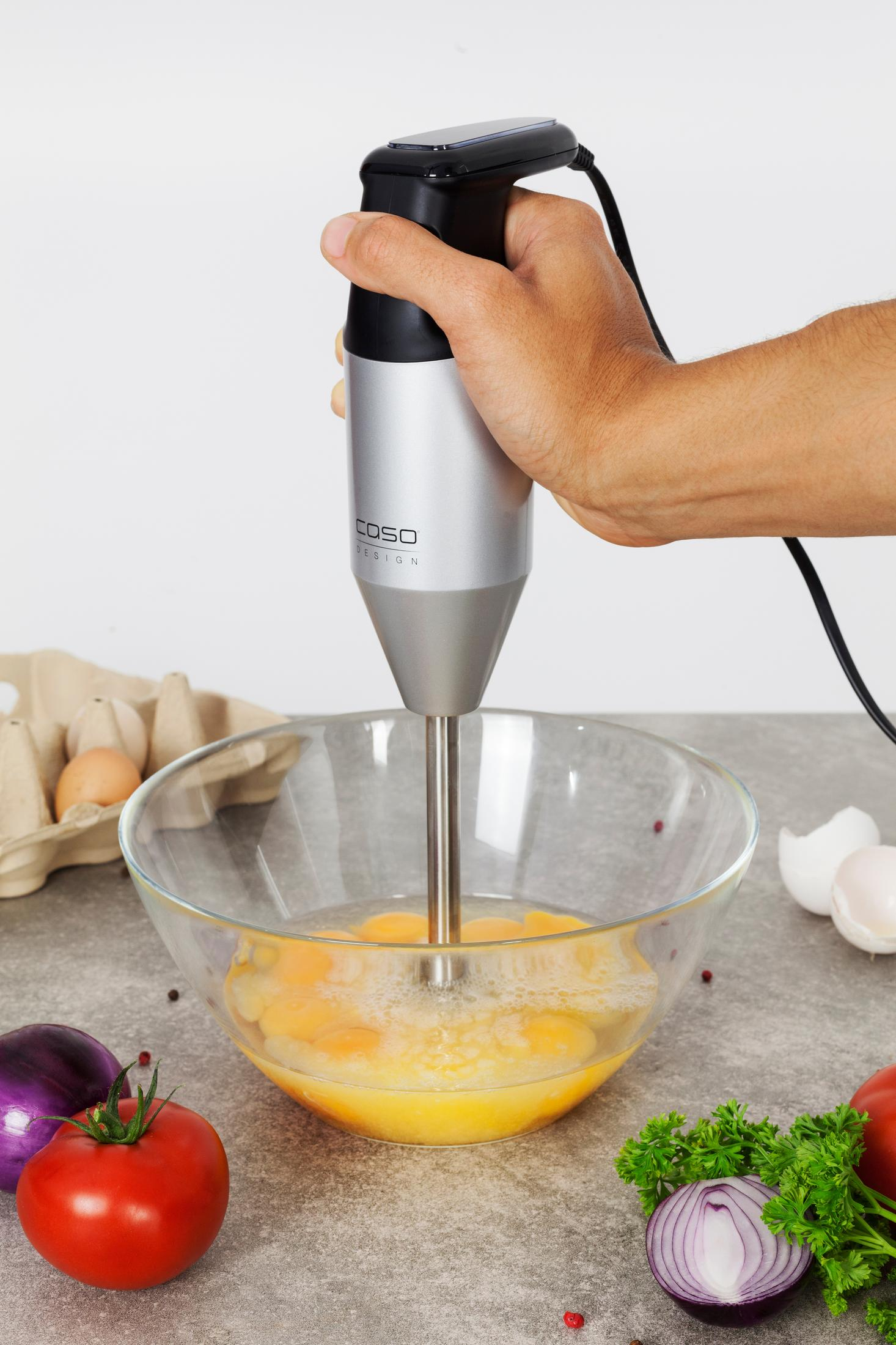 Mano mezclando huevos en un bol de cristal con una batidora de mano.