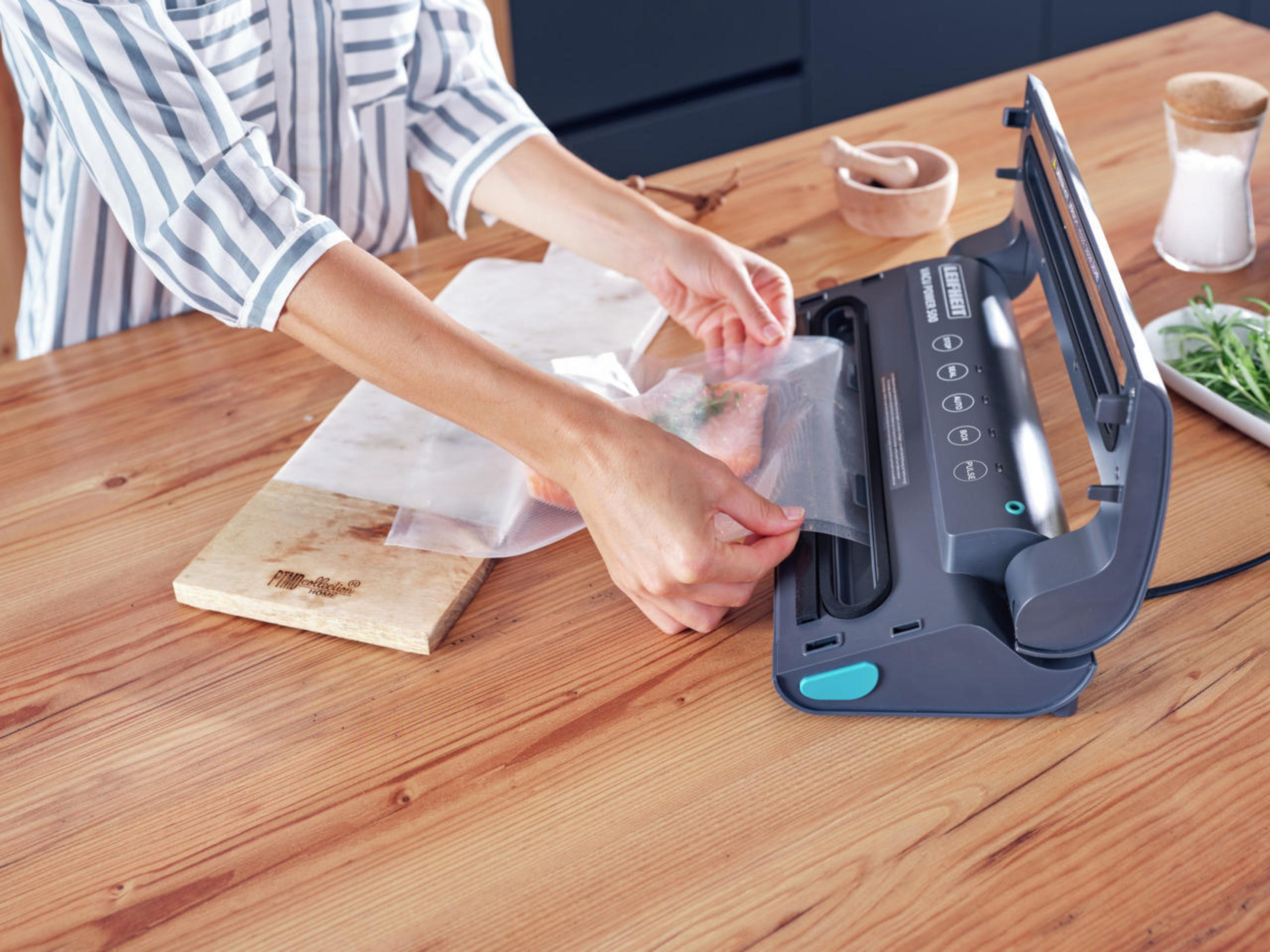 Eine Person versiegelt einen Beutel mit Essen mit einem Vakuumierer auf einem Holztisch.