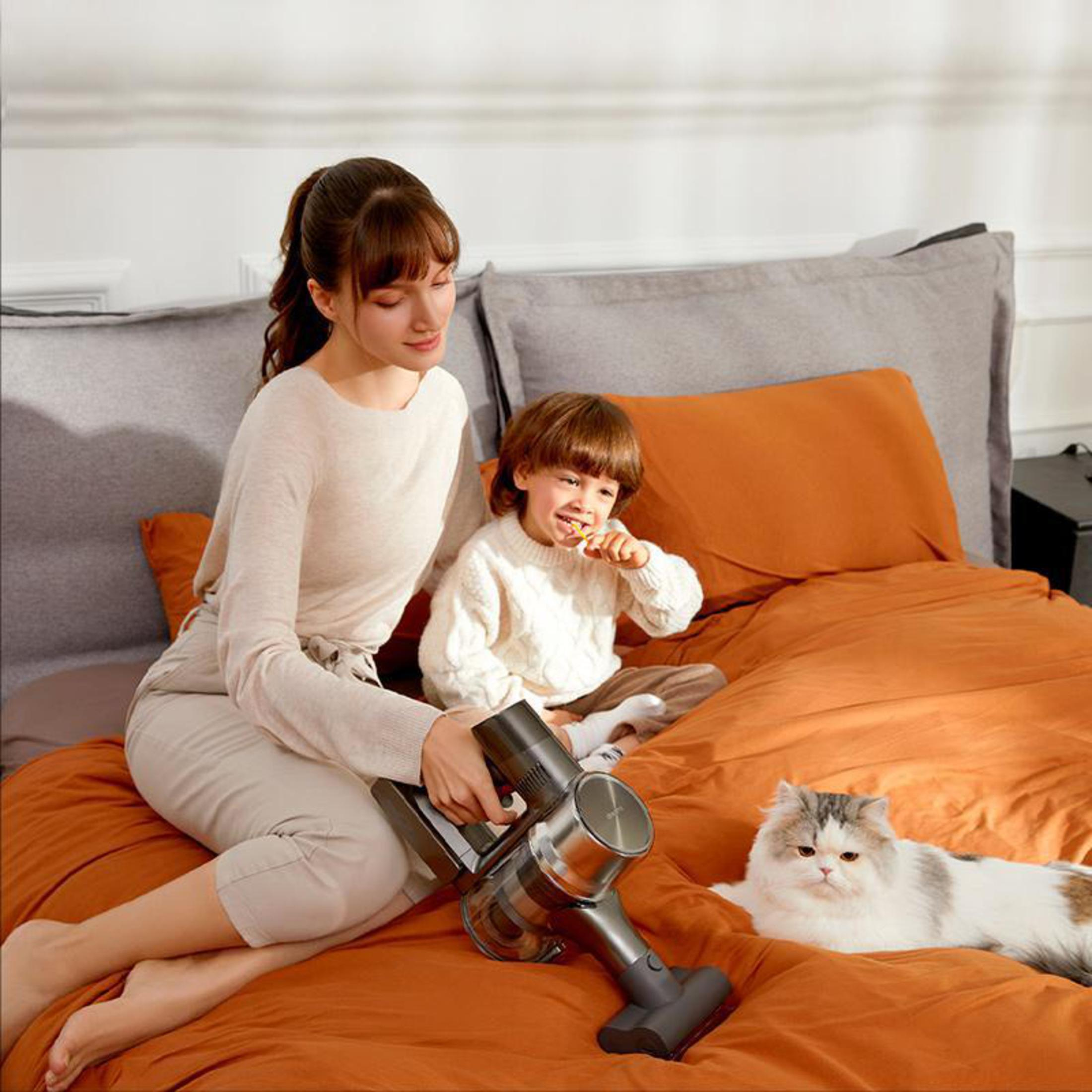 Eine Frau saugt das Bett ab, während ein Kind Zähne putzt und eine Katze anwesend ist.