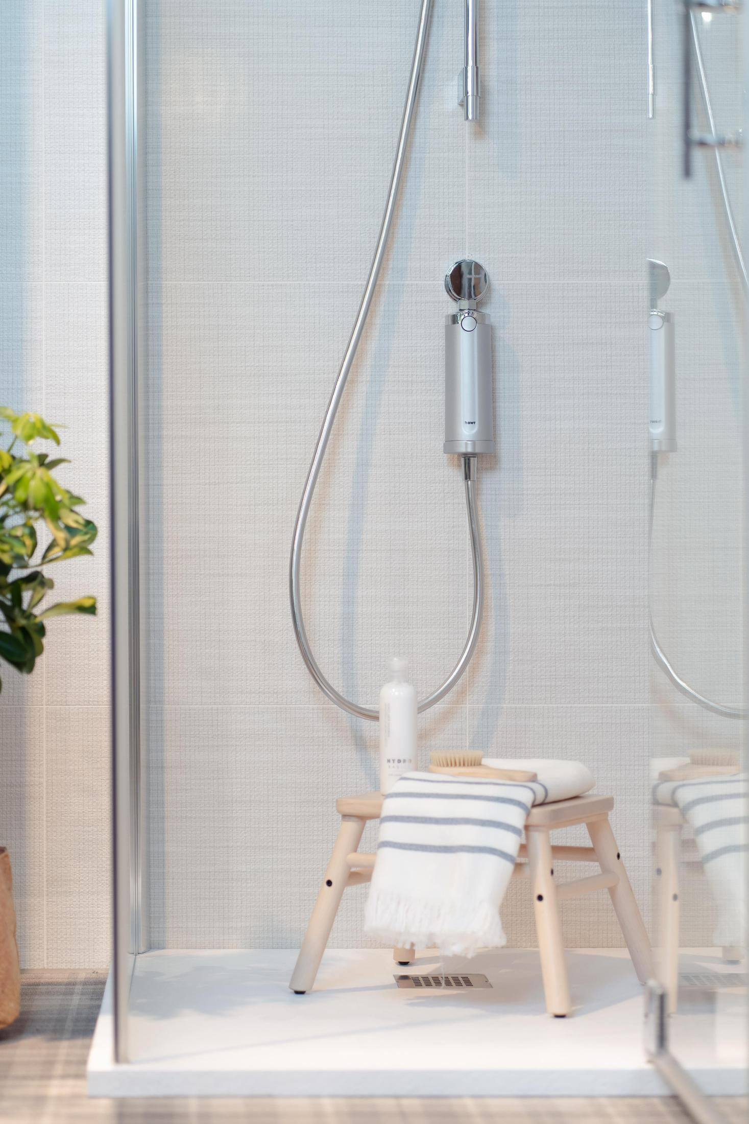 Kabina prysznicowa z drewnianym stołkiem, prysznicem i rośliną w rogu.