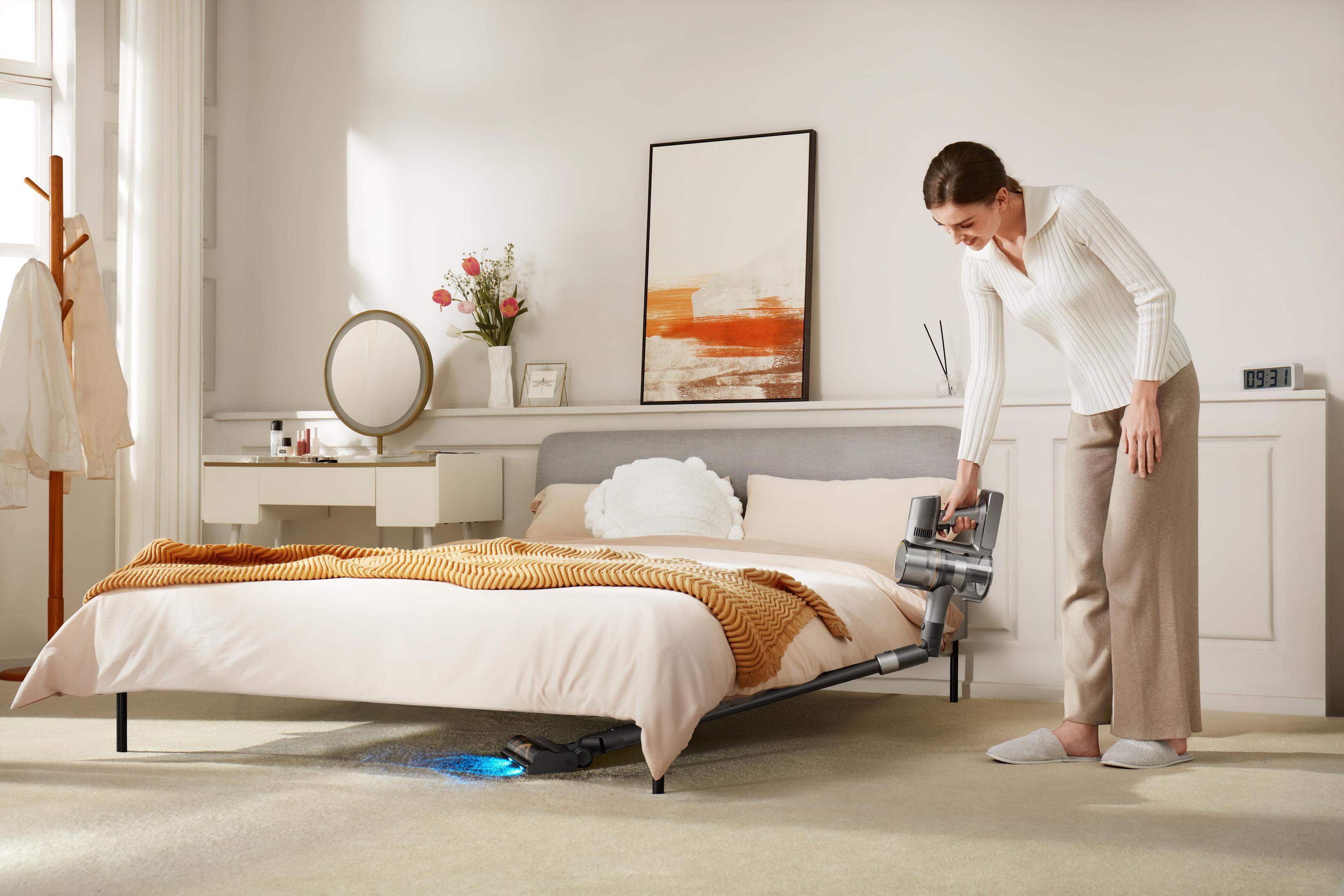 Frau, die ein Schlafzimmer mit einem kabellosen Staubsauger saugt. Bett, Spiegel und Kommode sichtbar.