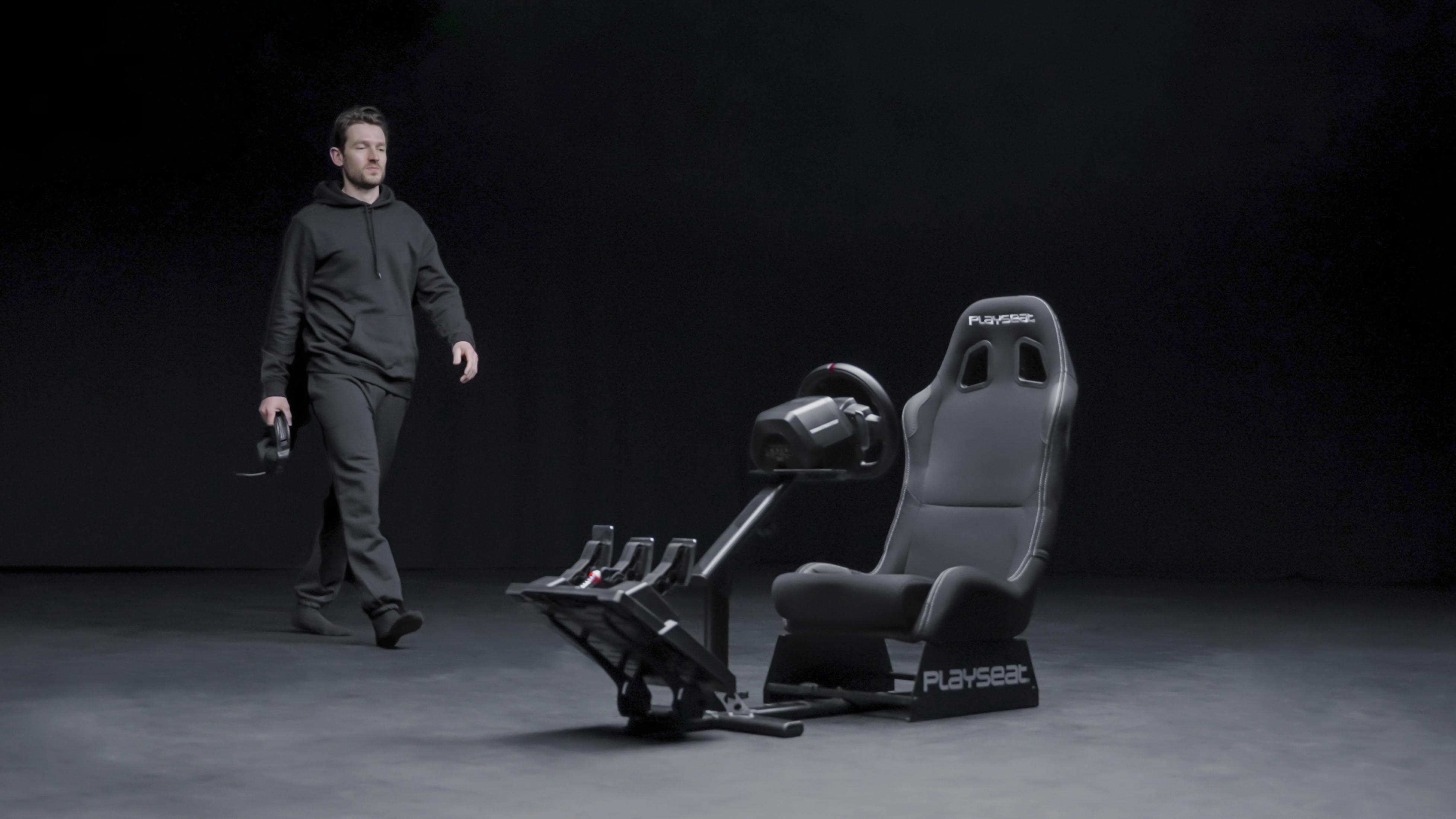 Mężczyzna idący w stronę fotela symulatora wyścigów, trzymający kontroler, ciemne tło.