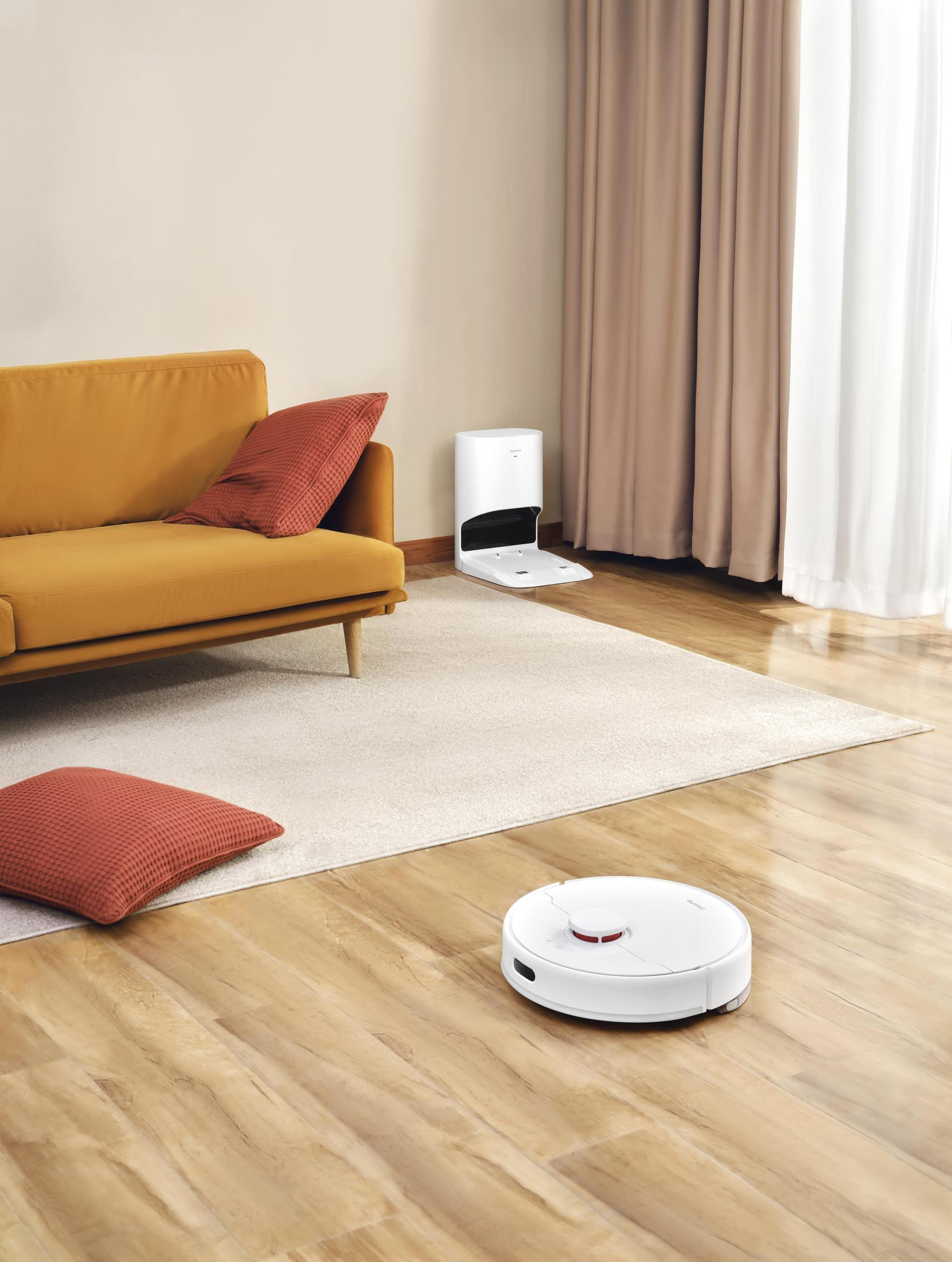 Una aspiradora robot blanca en una habitación, con una estación de carga en el fondo.