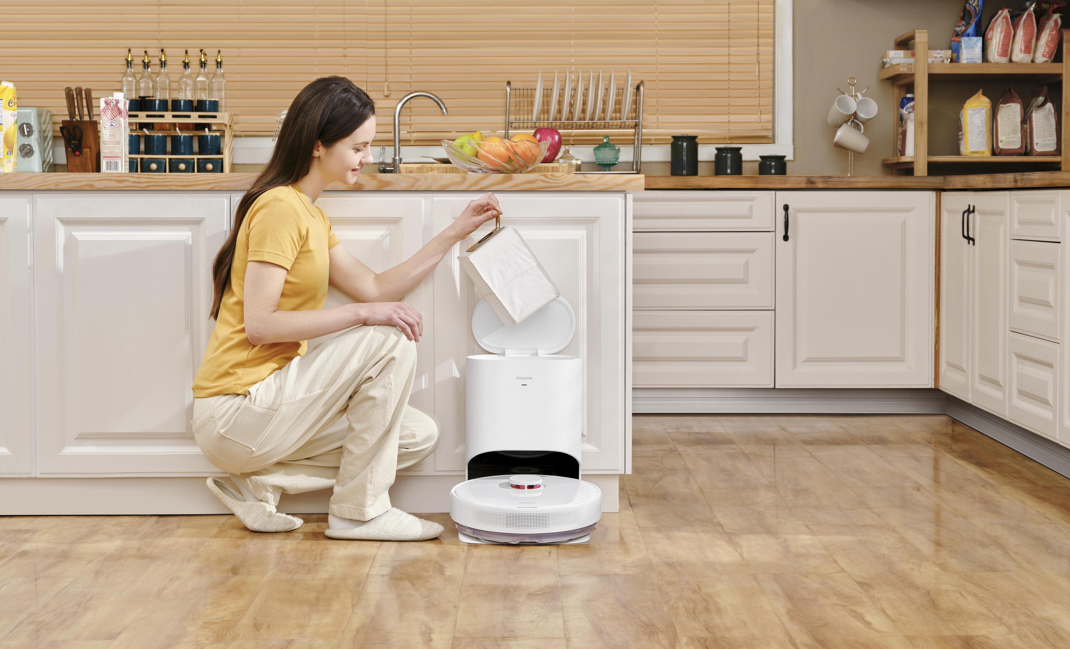 Una mujer poniendo una bolsa en una aspiradora robot. Fondo de cocina con gabinetes blancos y encimeras de madera.