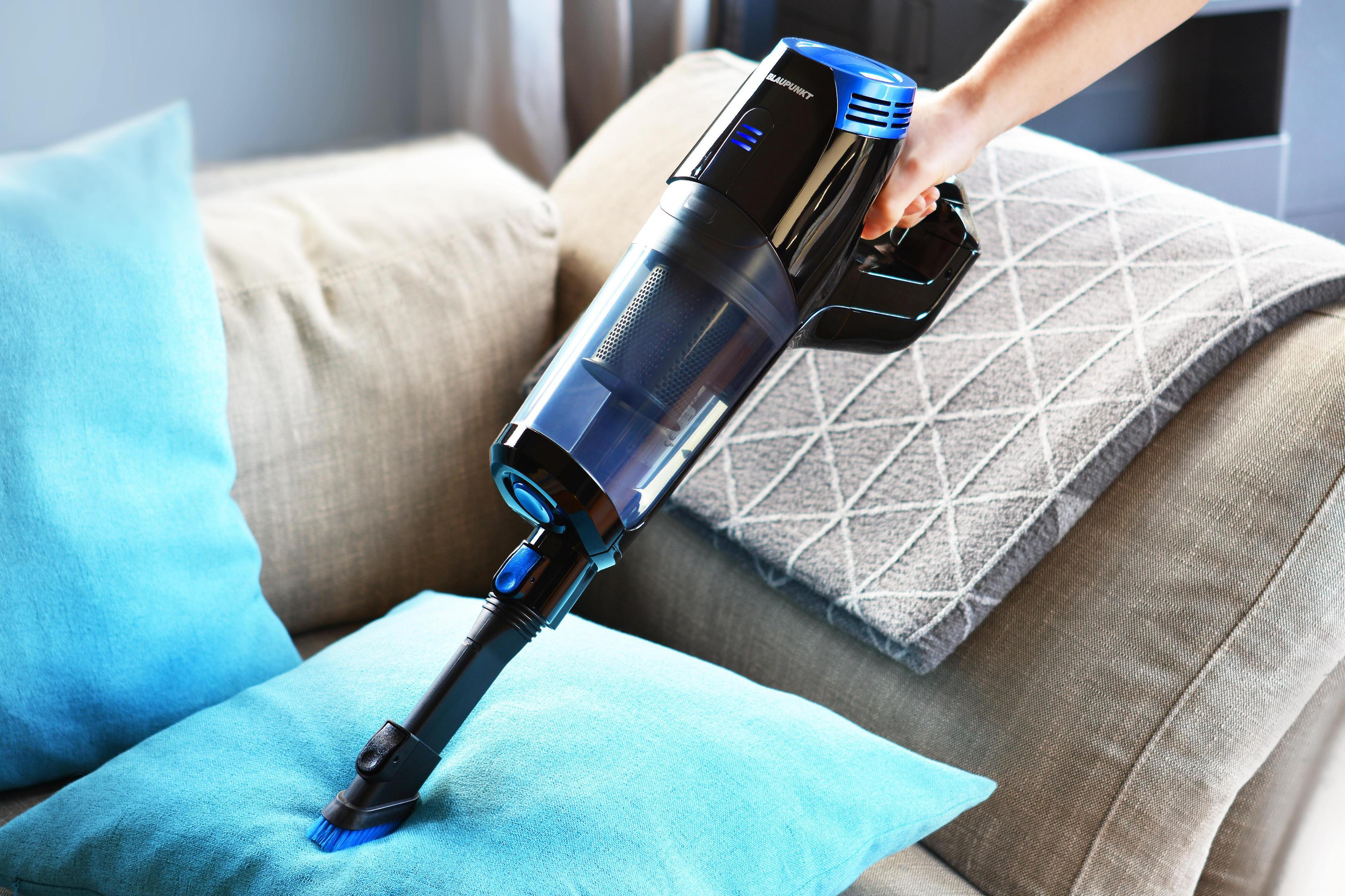 Osoba używająca czarno-niebieskiego bezprzewodowego odkurzacza do czyszczenia niebieskiej poduszki na sofie.
