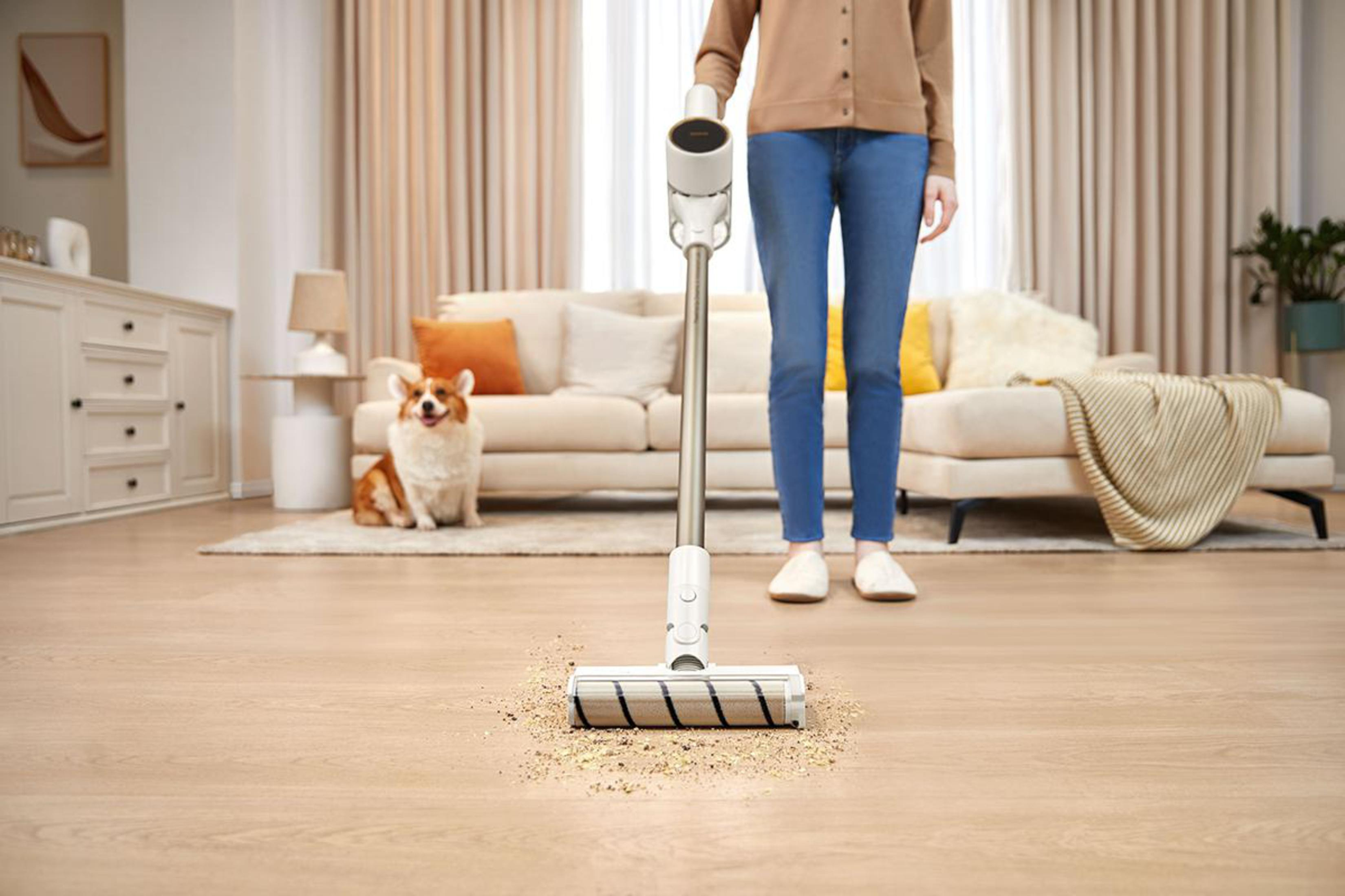 Una mujer que aspira un piso de madera. Un perro está sentado en el sofá, mirando la aspiradora.