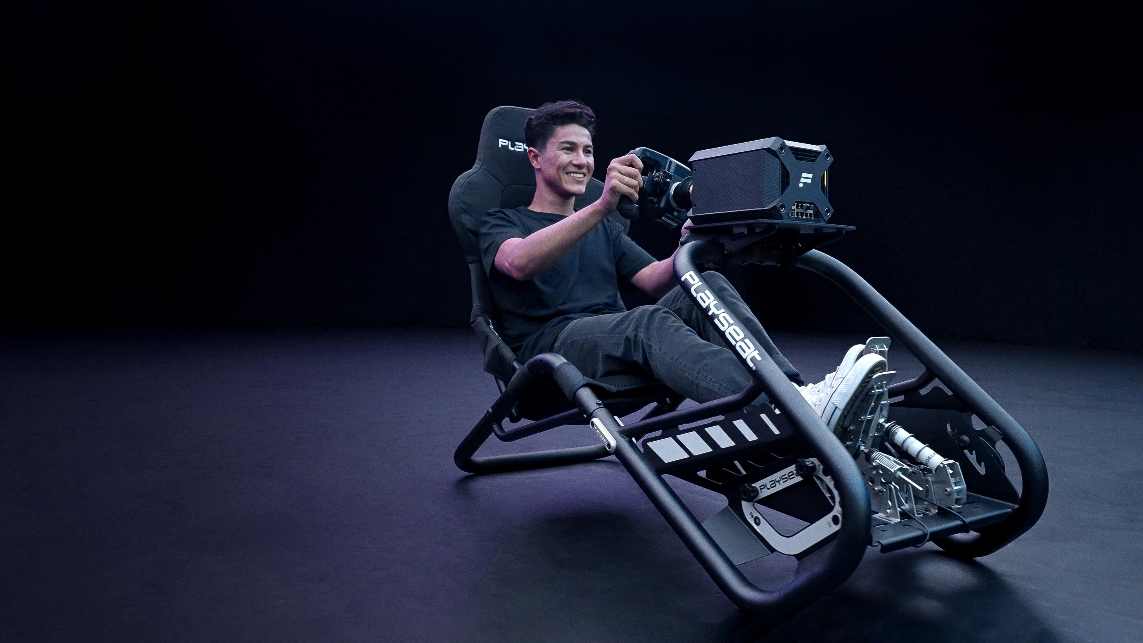 Mężczyzna siedzi na fotelu samochodu wyścigowego, trzymając kierownicę. Tło jest ciemne.