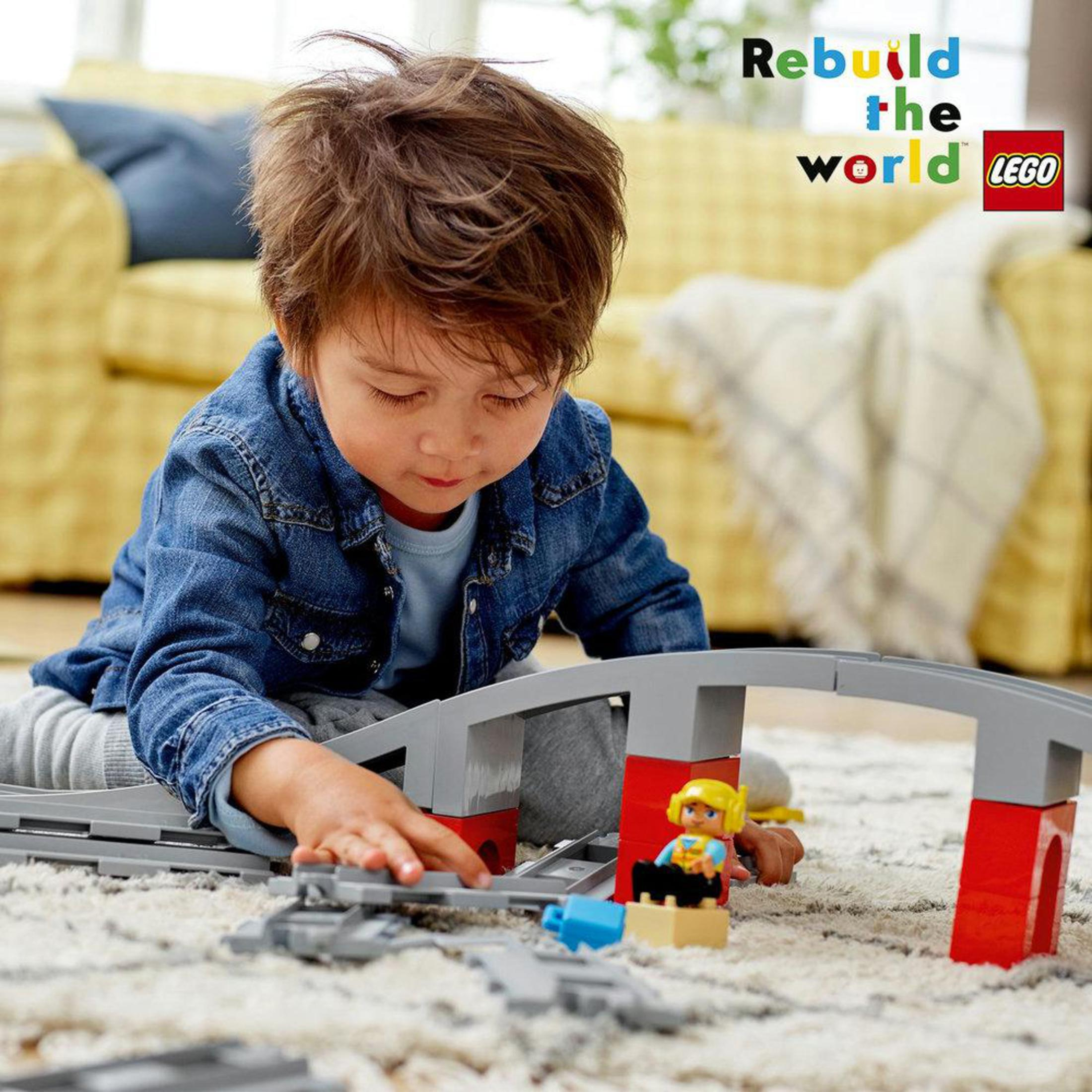 Bambino che gioca con un treno LEGO. Binari rossi e grigi. Figure LEGO presenti.