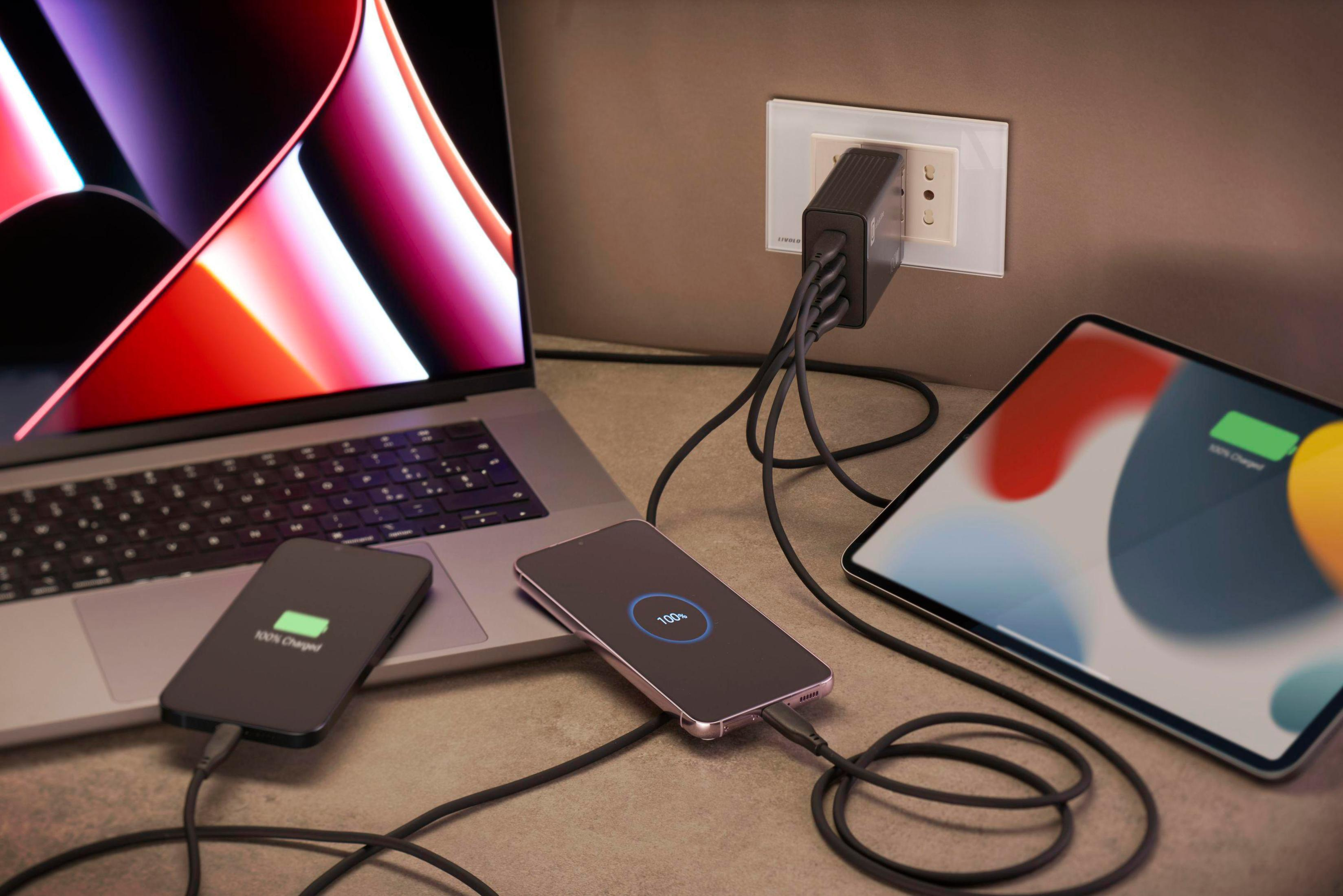 Laptop, Telefon und Tablet laden mit mehreren Kabeln. Der Hintergrund ist eine Wand und eine Steckdose.
