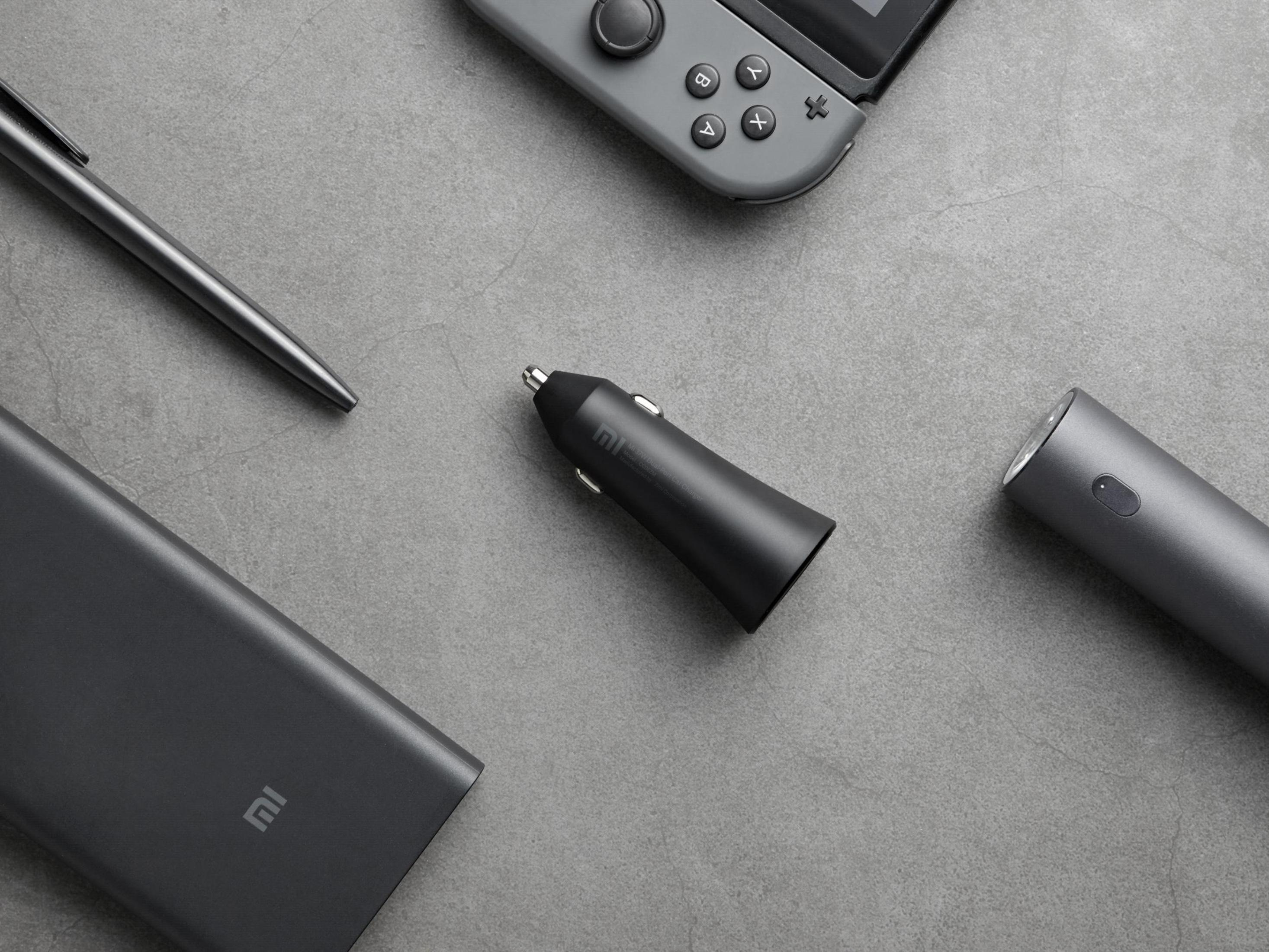 Czarna ładowarka samochodowa, power bank, długopis i latarka na szarej powierzchni. Widoczna jest również konsola do gier.