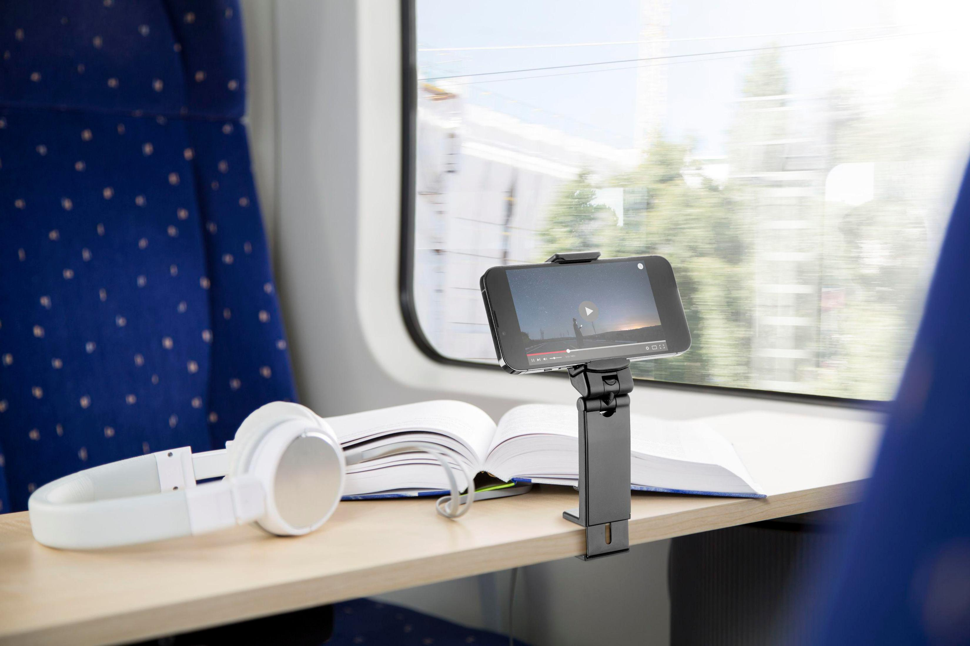 Un telefono che mostra video è montato su un tavolino in treno. Cuffie e un libro sono presenti.