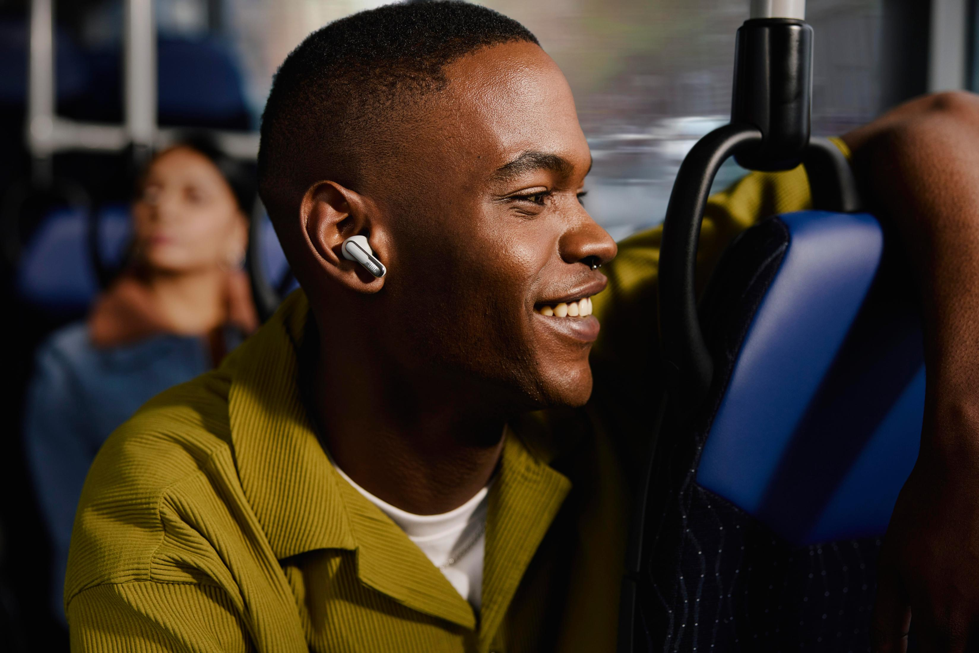 Un uomo con un auricolare nell'orecchio, seduto su un autobus. Sta sorridendo e guarda fuori dal finestrino.