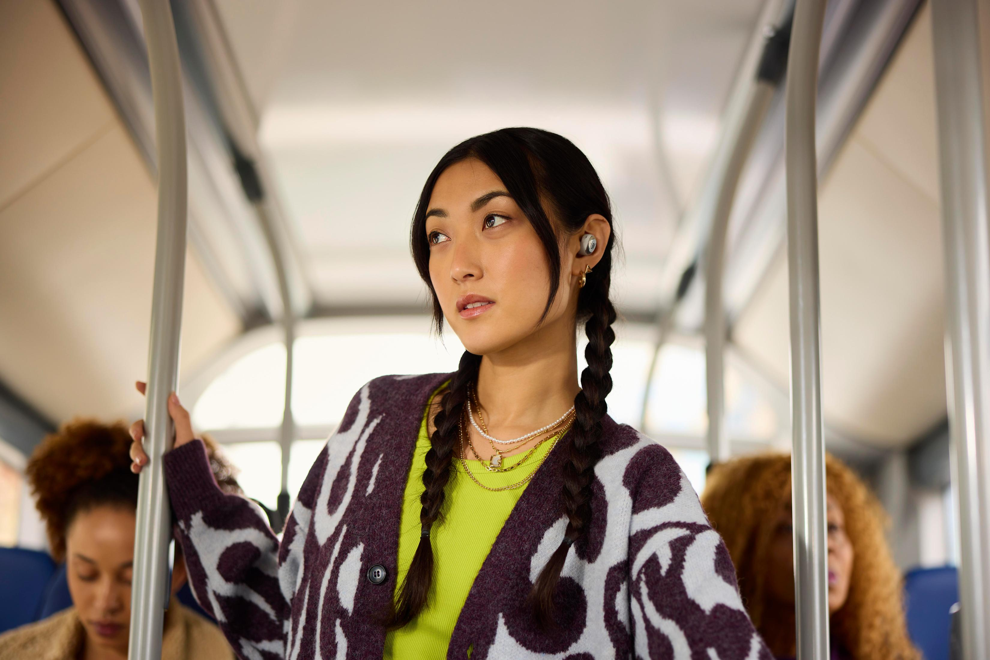 Donna in autobus con auricolari. Ha trecce, una giacca fantasia e sembra pensierosa.