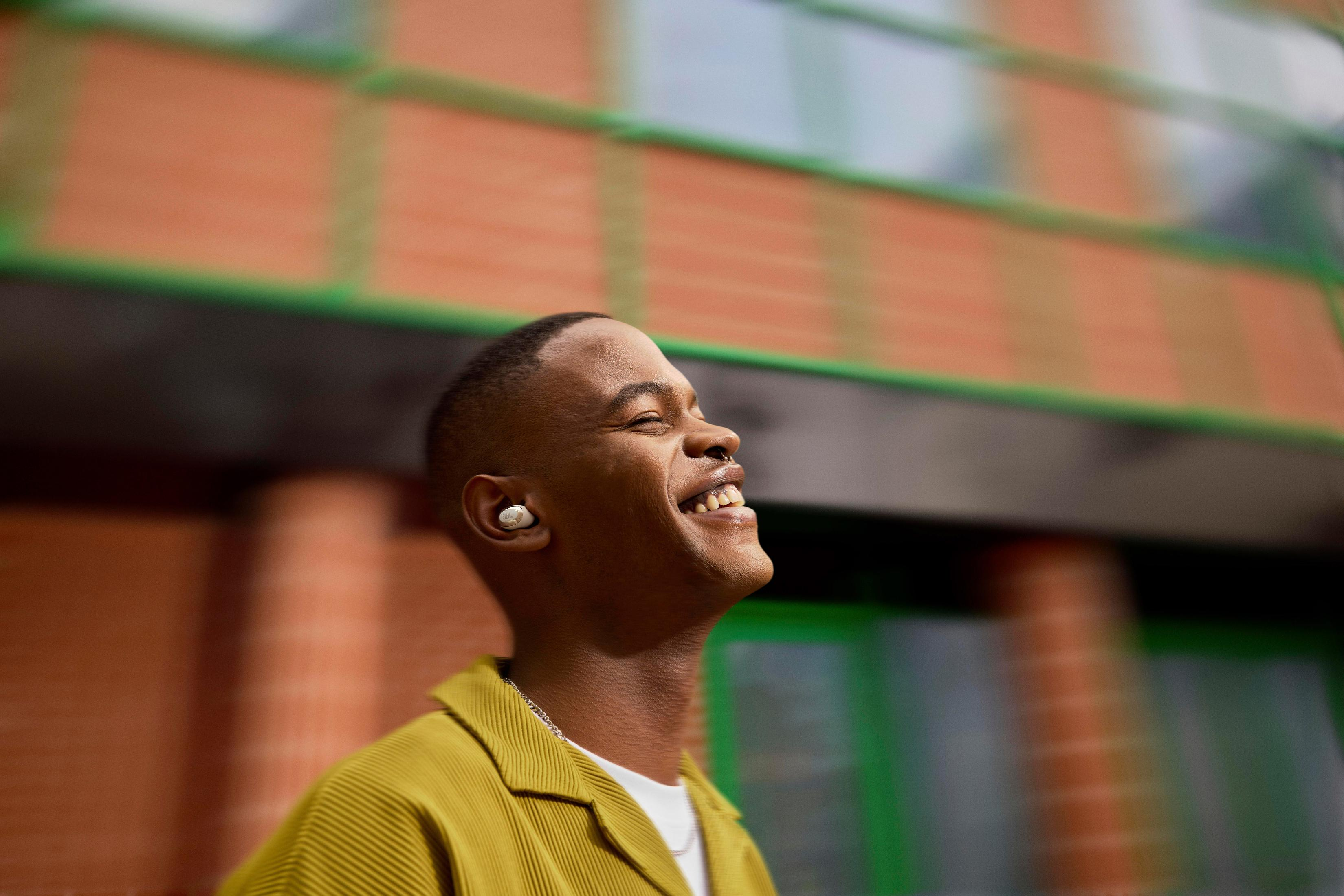 Un uomo nero sorridente con auricolari, con una camicia gialla, che guarda in alto.