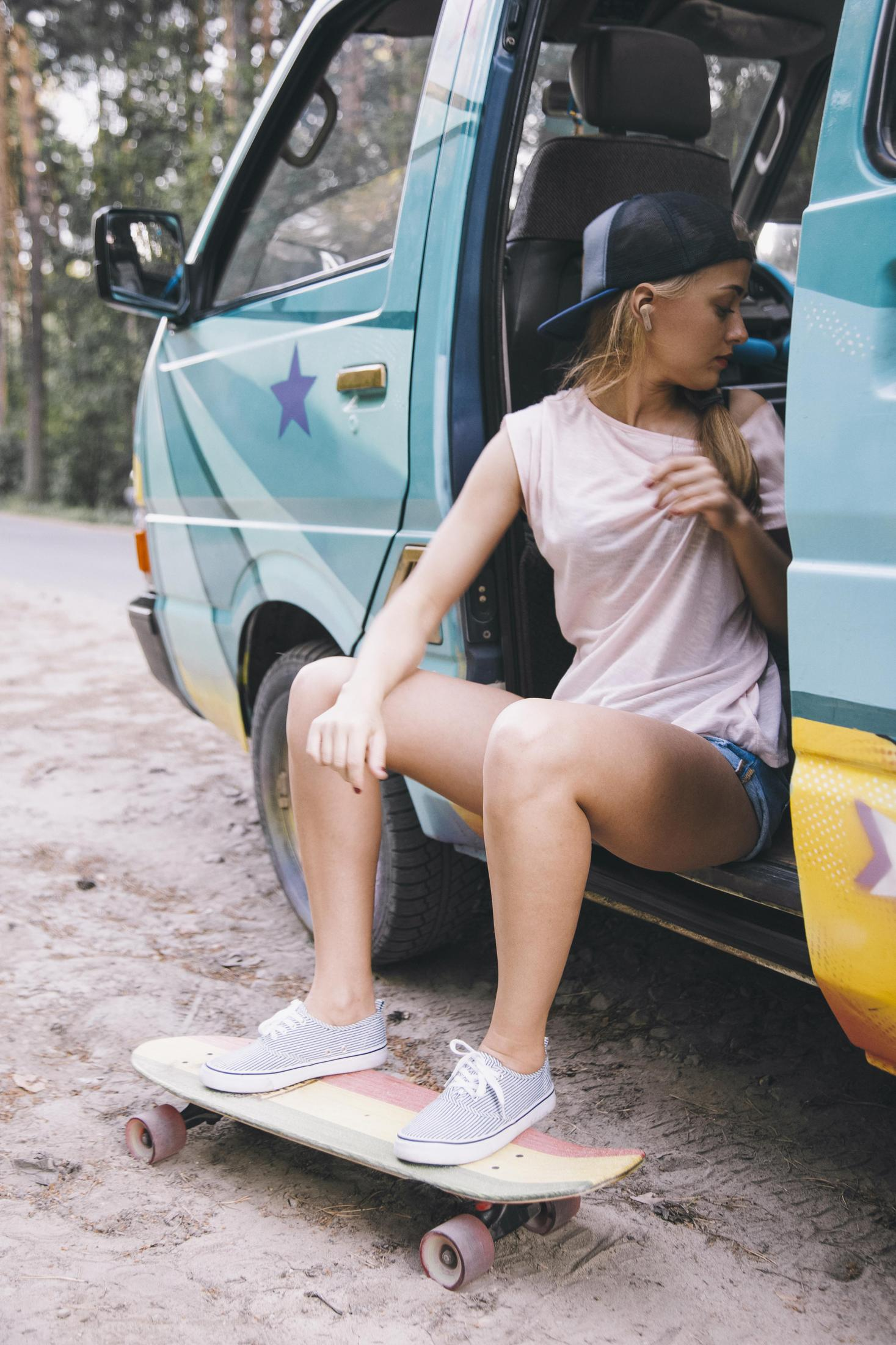 Eine Frau sitzt auf einem Skateboard neben einem türkisfarbenen Van. Sie trägt ein rosa Hemd und eine schwarze Kappe.