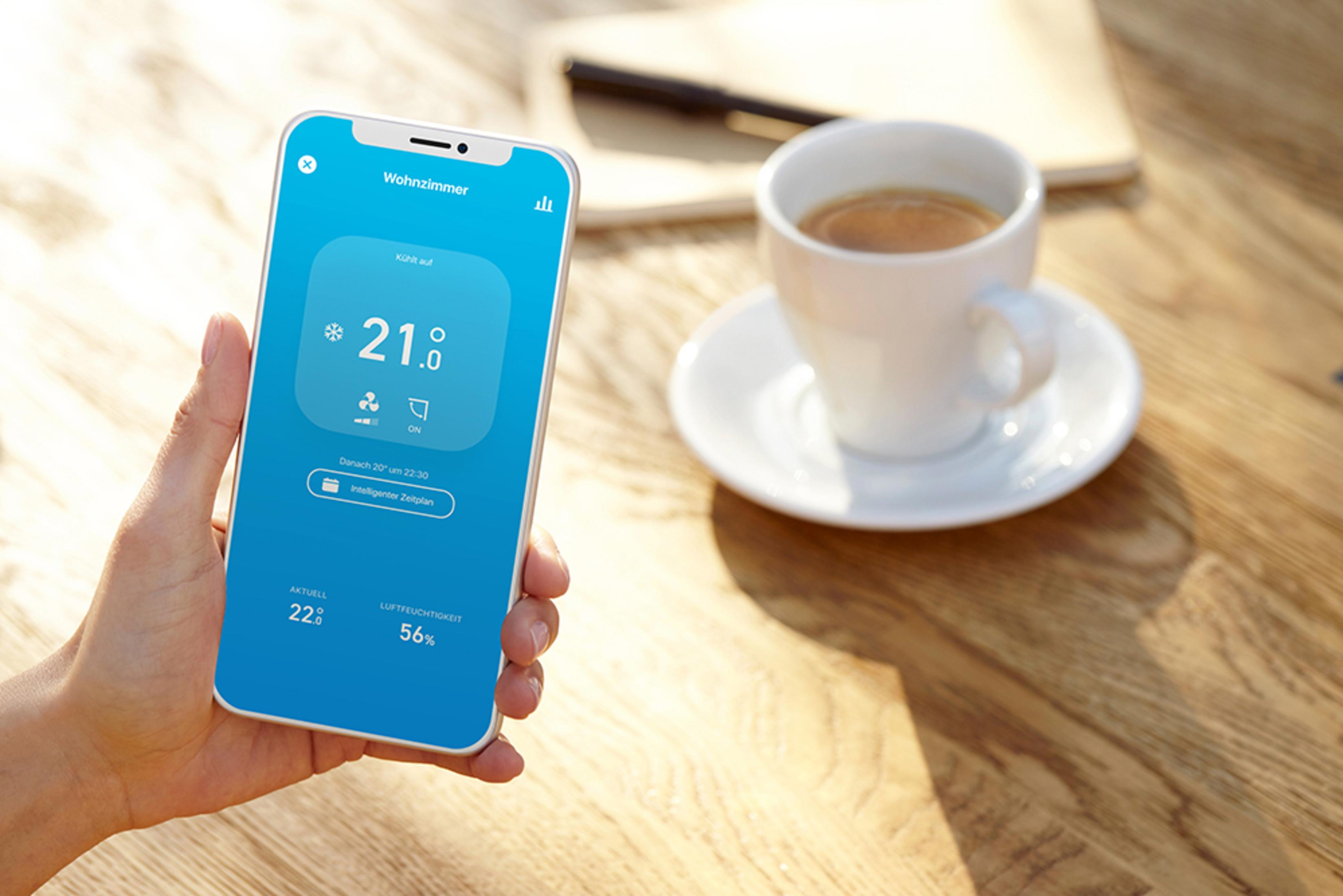 A person's hand holding a phone displaying a smart home app interface next to a coffee cup.