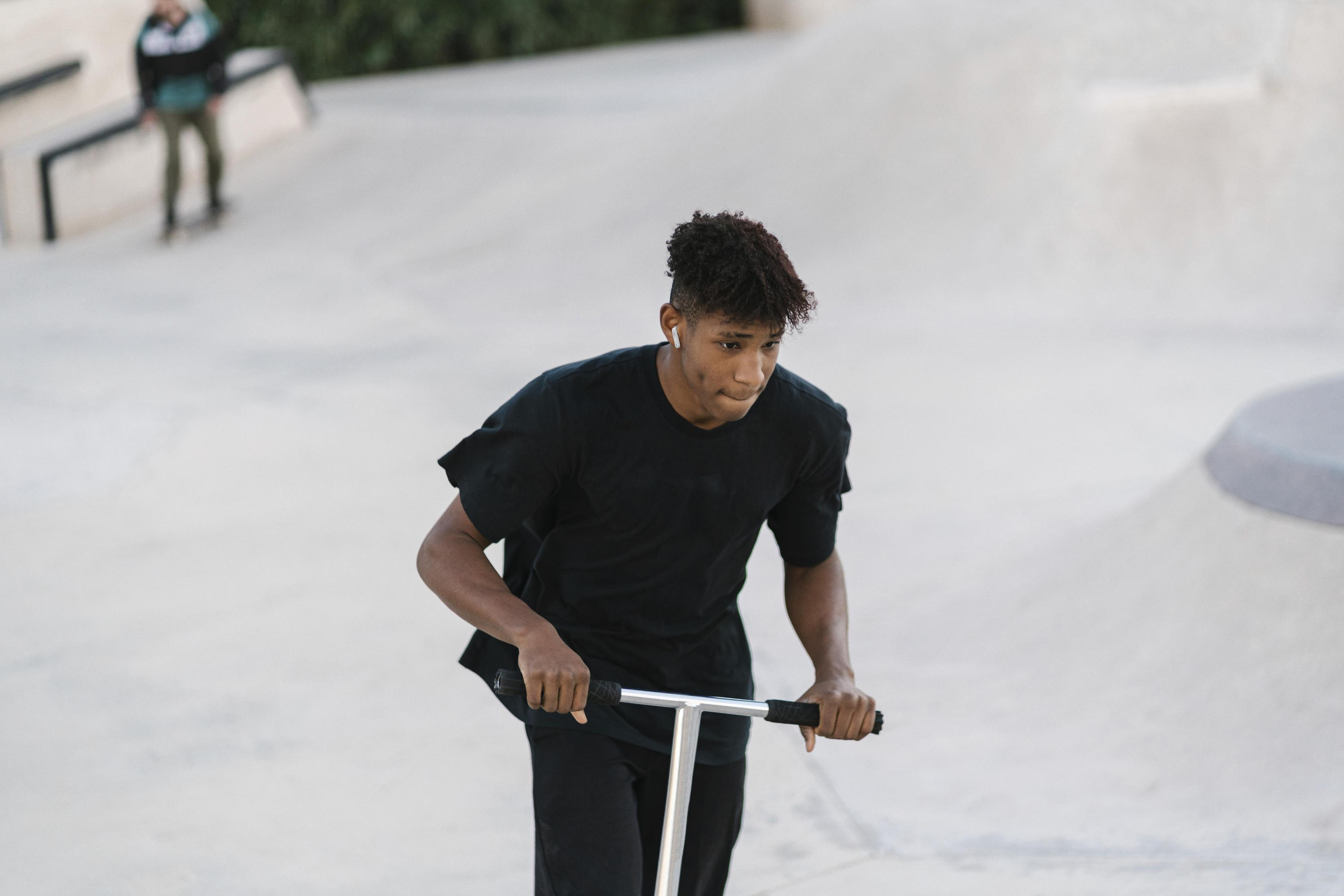 Junger Mann fährt mit einem Roller in einem Skatepark. Er trägt ein schwarzes Outfit.