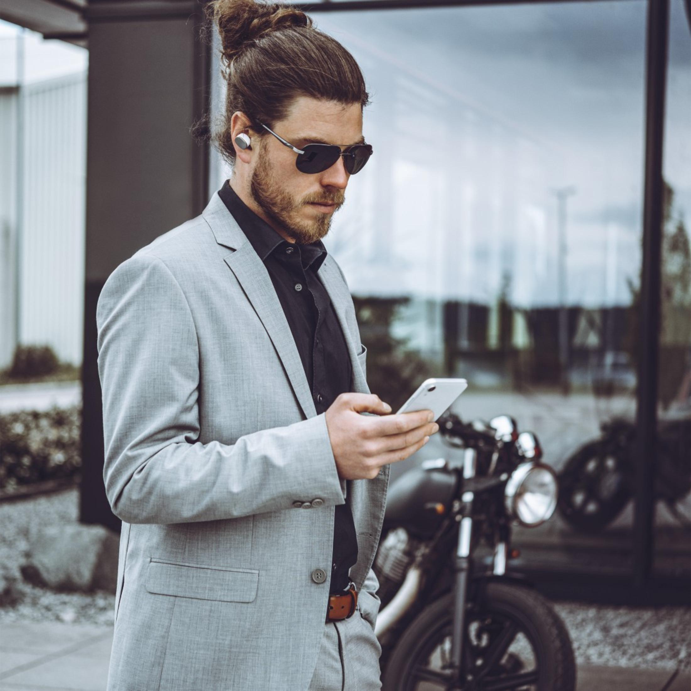 Ein Mann in grauem Anzug, Sonnenbrille und Ohrhörern benutzt ein Smartphone neben Motorrad.