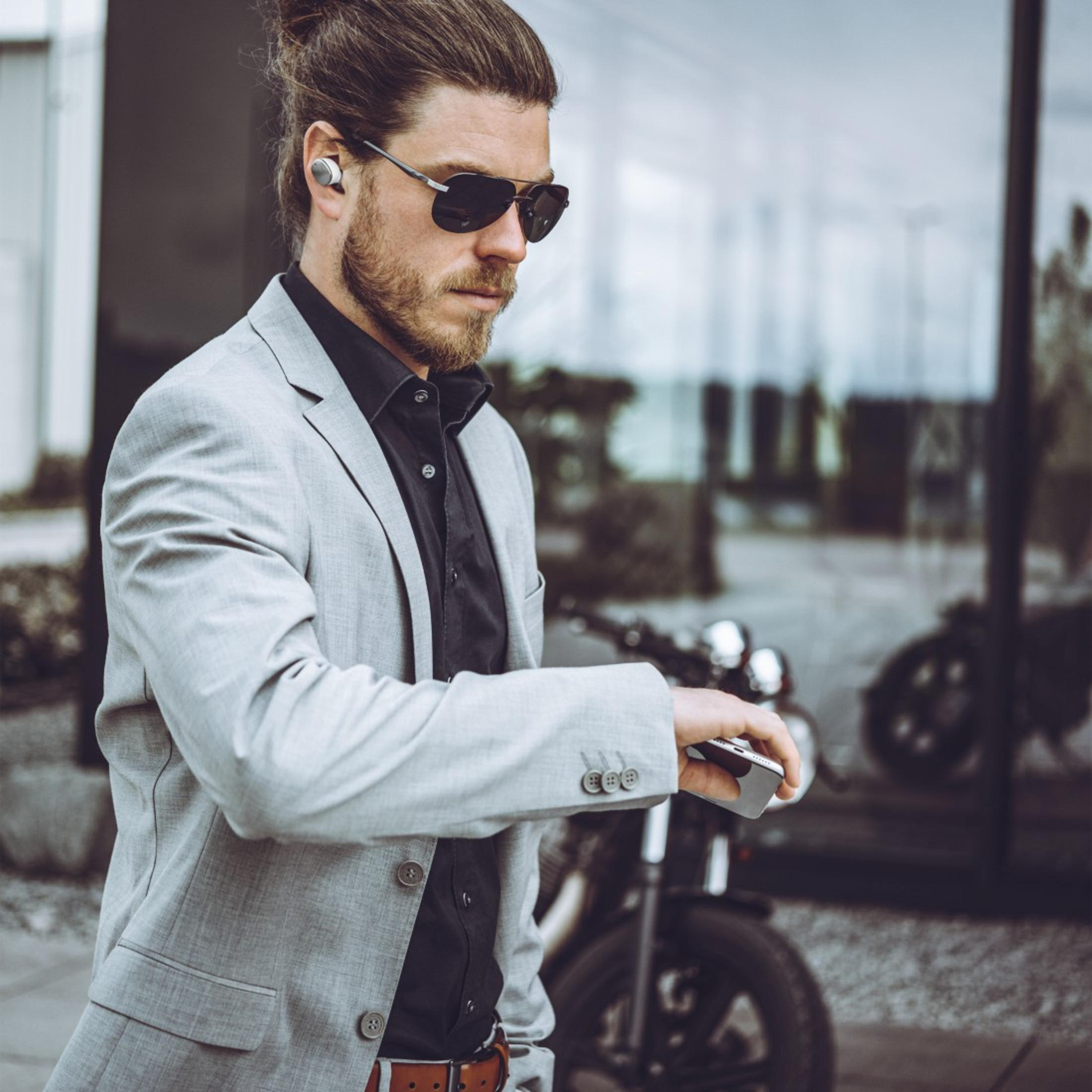 Ein Mann mit Sonnenbrille und grauem Anzug hält ein Smartphone, mit Ohrhörern in den Ohren.