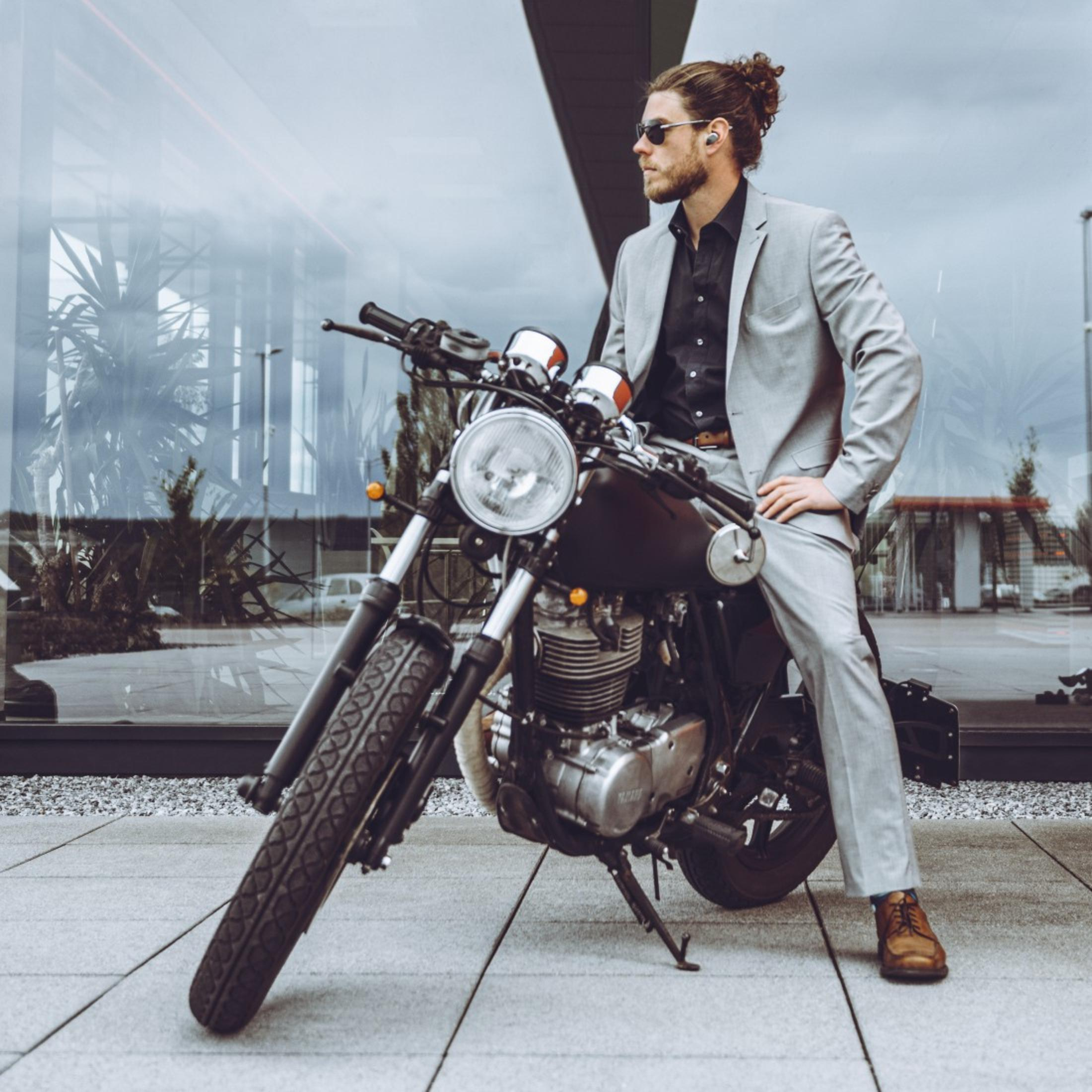 Ein Mann in einem grauen Anzug sitzt auf einem schwarzen Motorrad, mit Sonnenbrille und modernem Hintergrund.