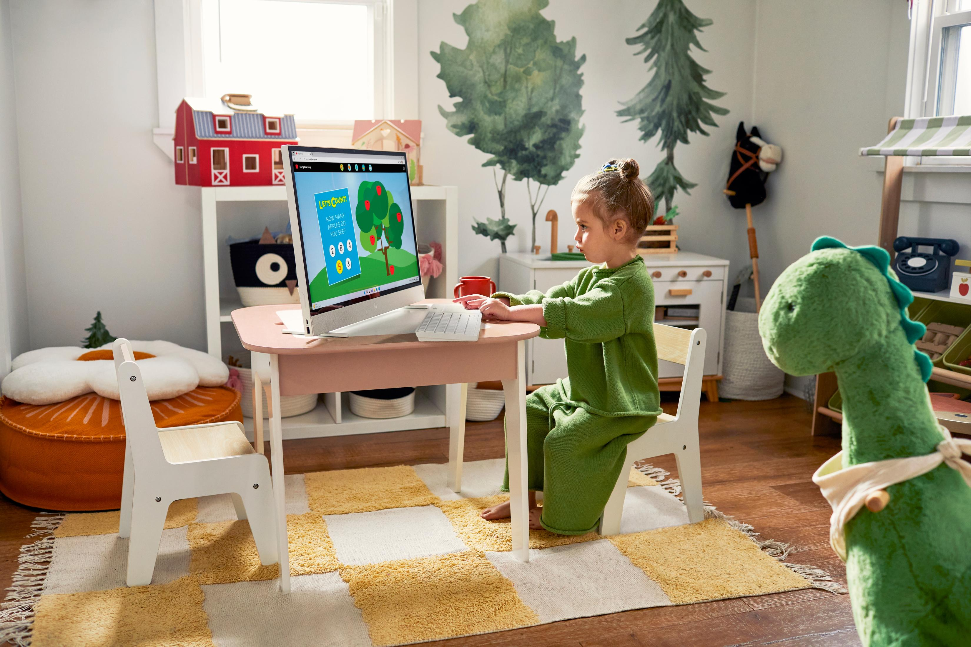 Dziewczynka w zielonym stroju siedzi przy różowym biurku, patrząc na ekran komputera z grą.