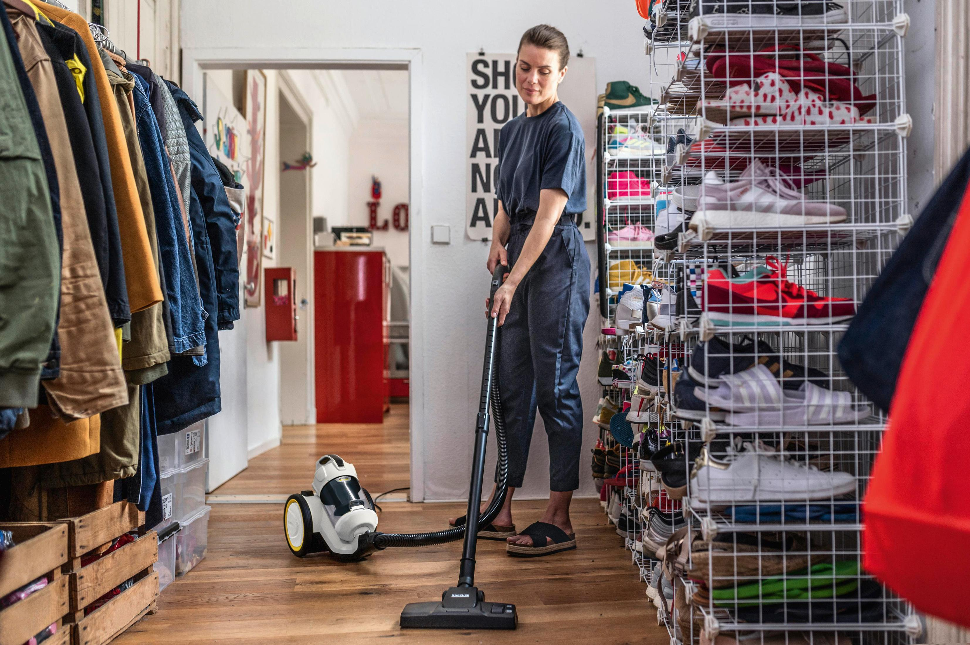 Kobieta odkurza drewnianą podłogę w korytarzu. Jest odkurzacz i buty na półkach.