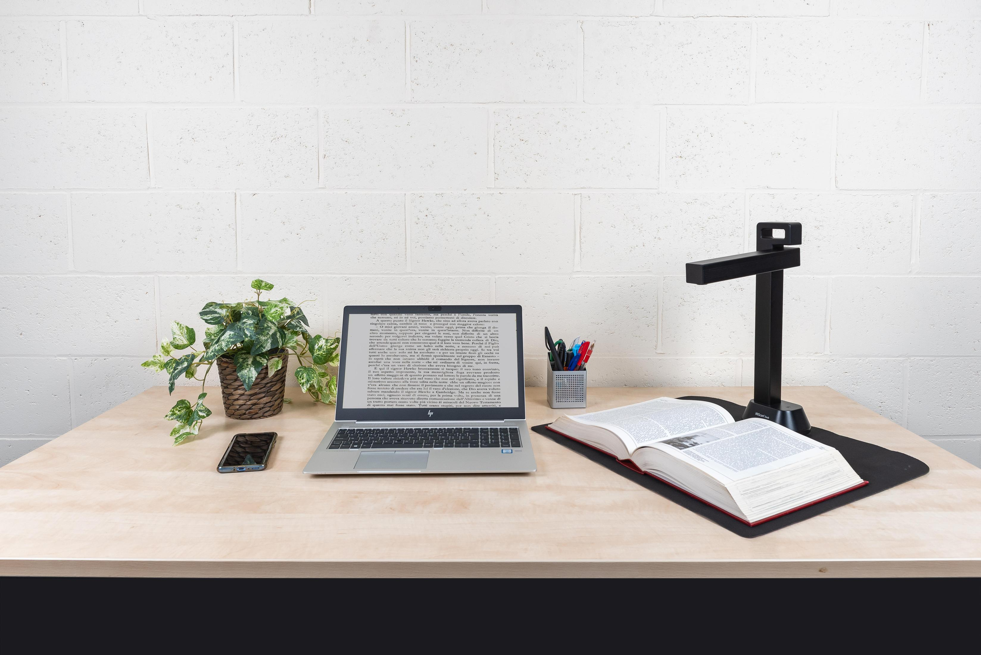 Laptop, książka, telefon i skaner znajdują się na drewnianym biurku. W tle roślina.